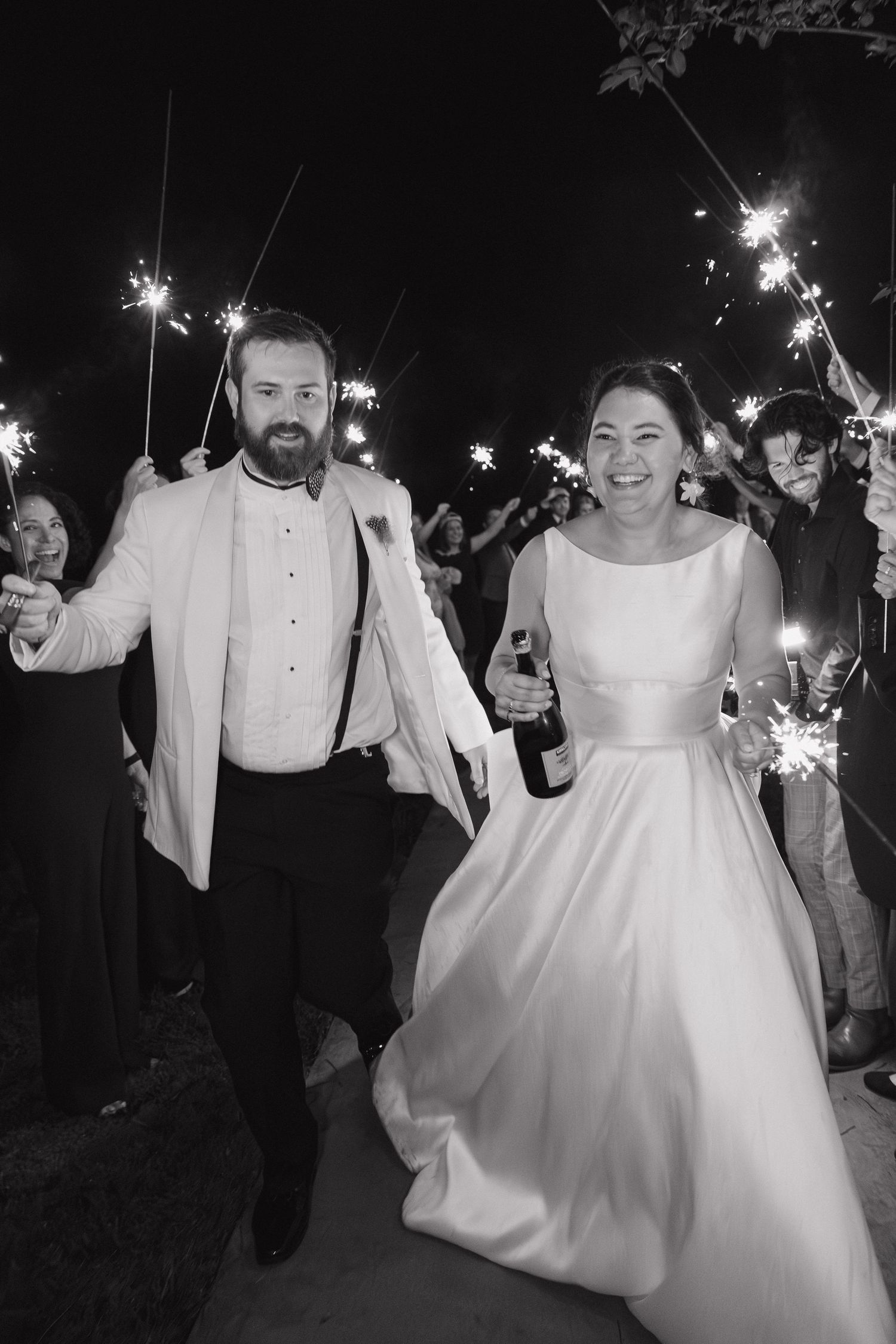 Joyful bride and groom walk through sparkler send-off holding champagne bottle in classic black and white wedding photo.