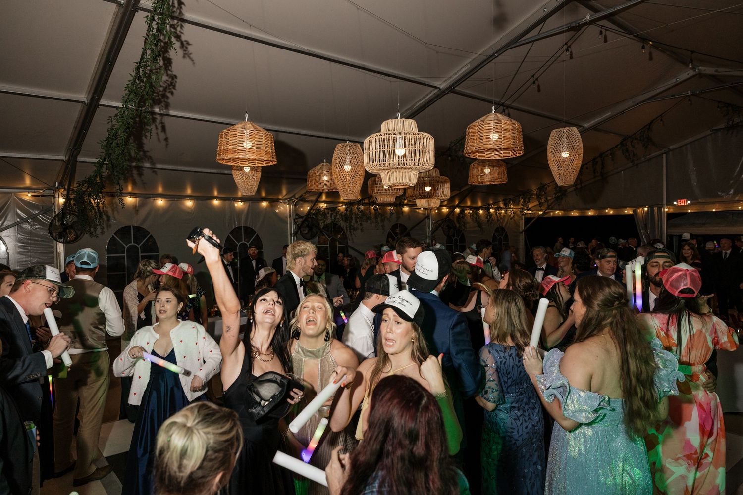 Lively wedding reception dance floor packed with guests under wicker chandelier lights in a white tent venue.