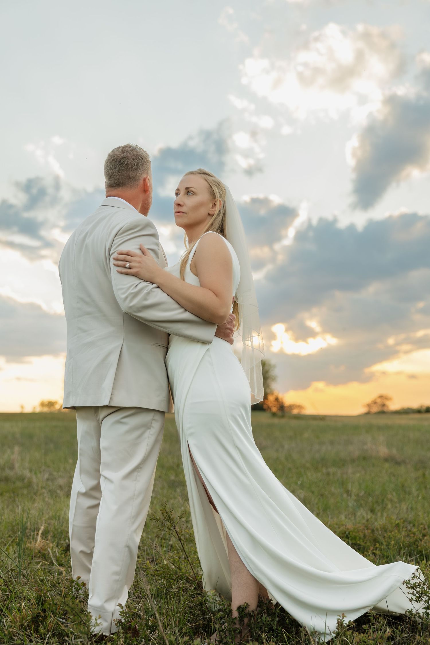 Bride and groom embrace in a grassy field with a dramatic sunset sky.