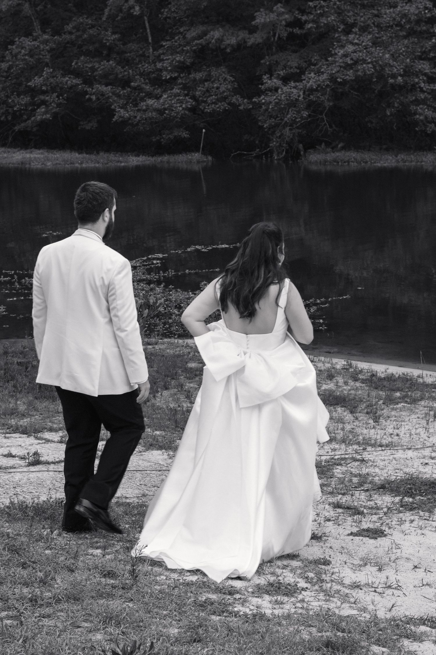 Black and white photo of bride and groom walking toward a peaceful lake on their wedding day.
