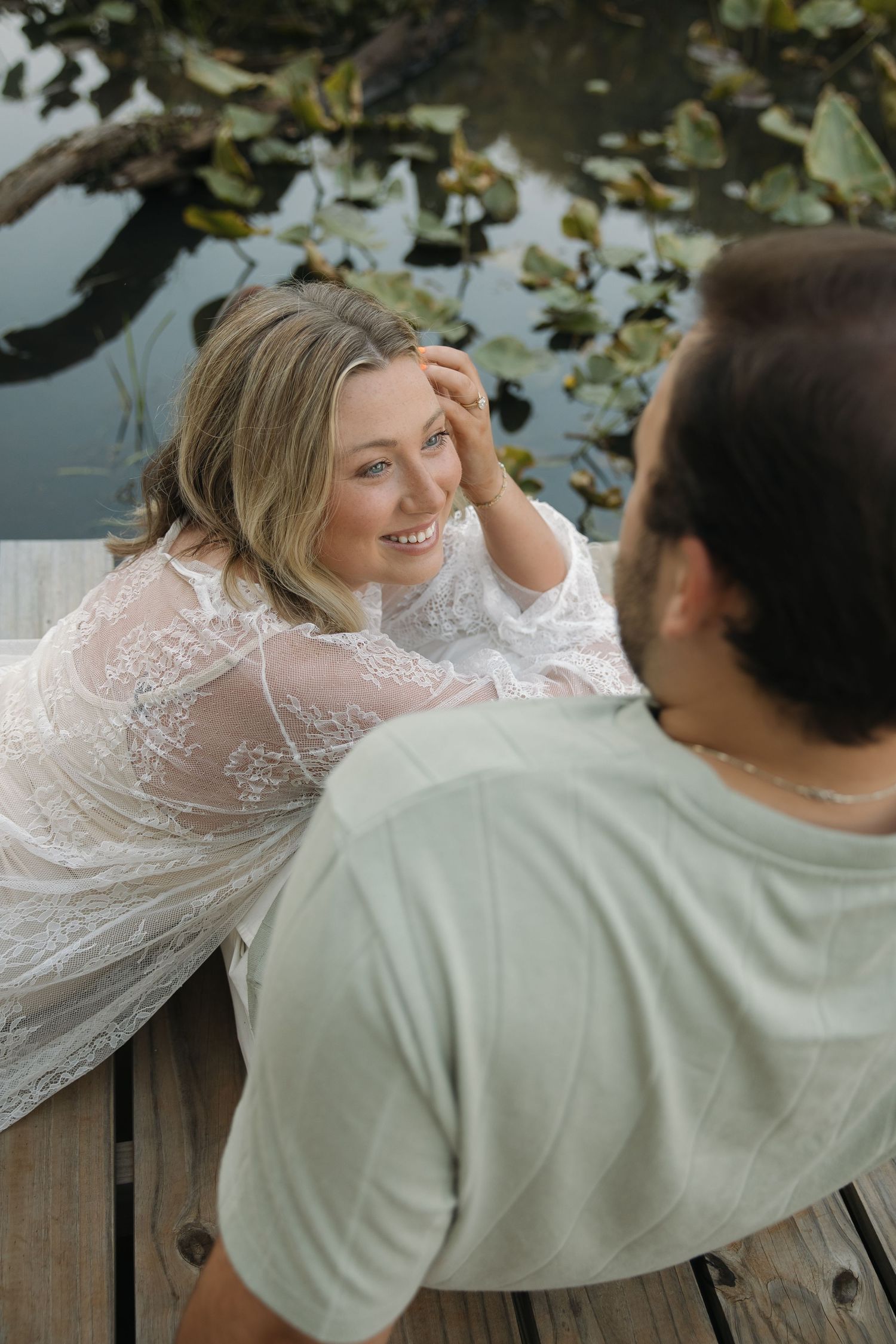 Smiling woman in white lace dress lying on a dock gazing up at her partner near a lily pad pond.