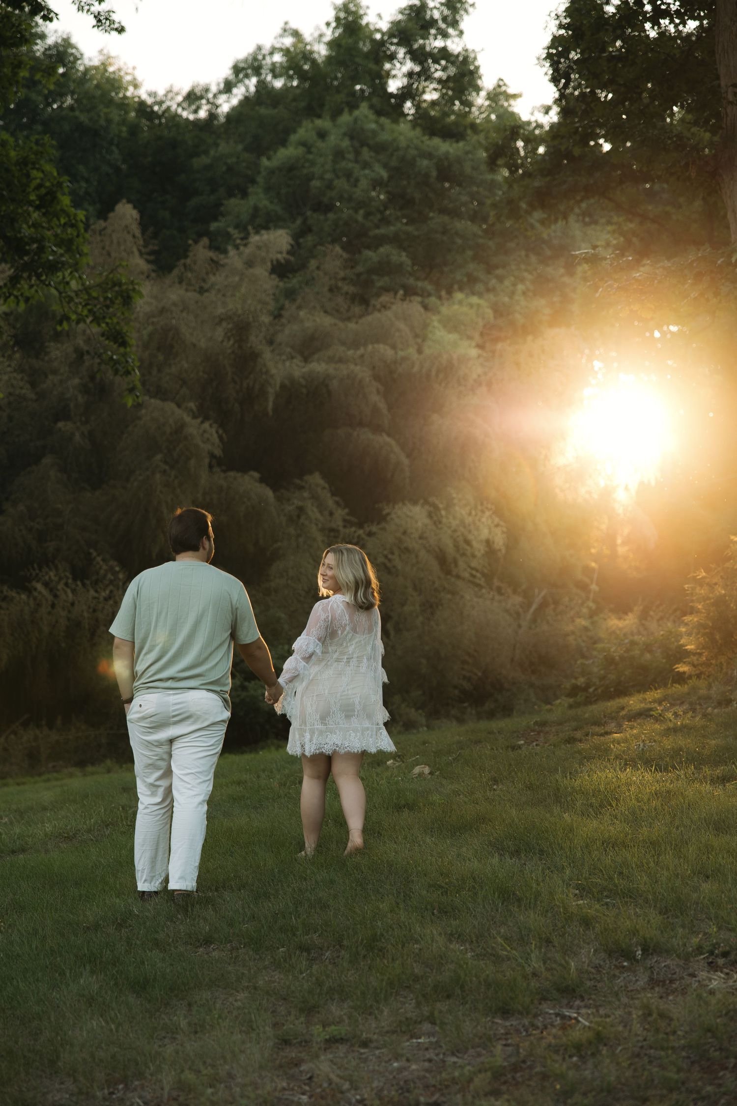 Couple holding hands walking toward golden sunset in lush green park, woman in white lace dress, man in mint shirt.