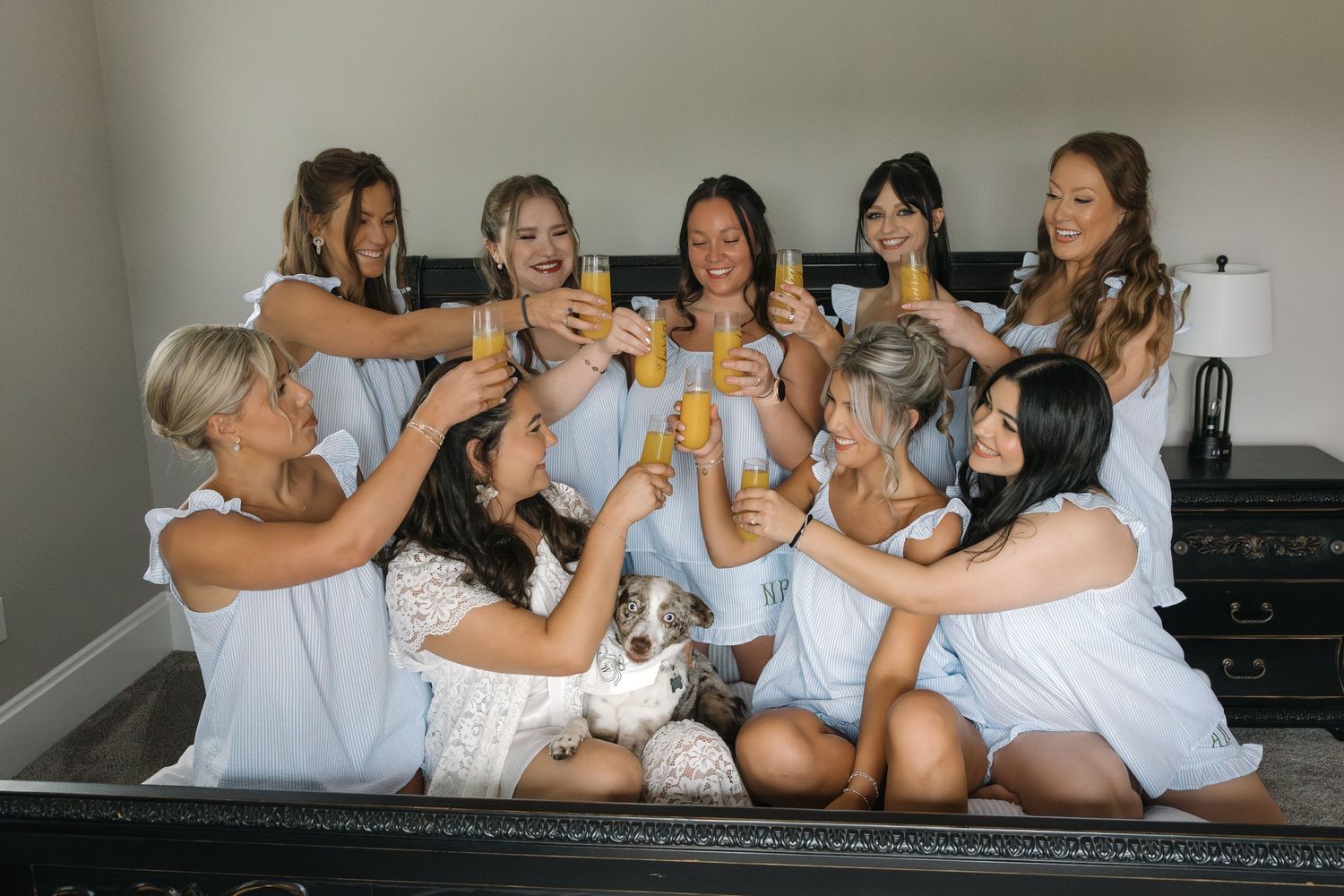 Bridesmaids in white robes raising mimosa glasses in a cheerful toast with a puppy.
