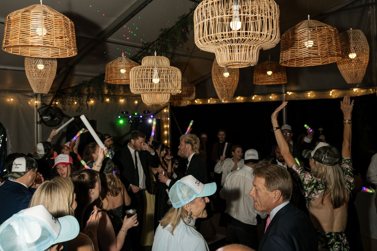 Energetic wedding reception dance floor with guests holding glow sticks under wicker pendant lights in a tent.