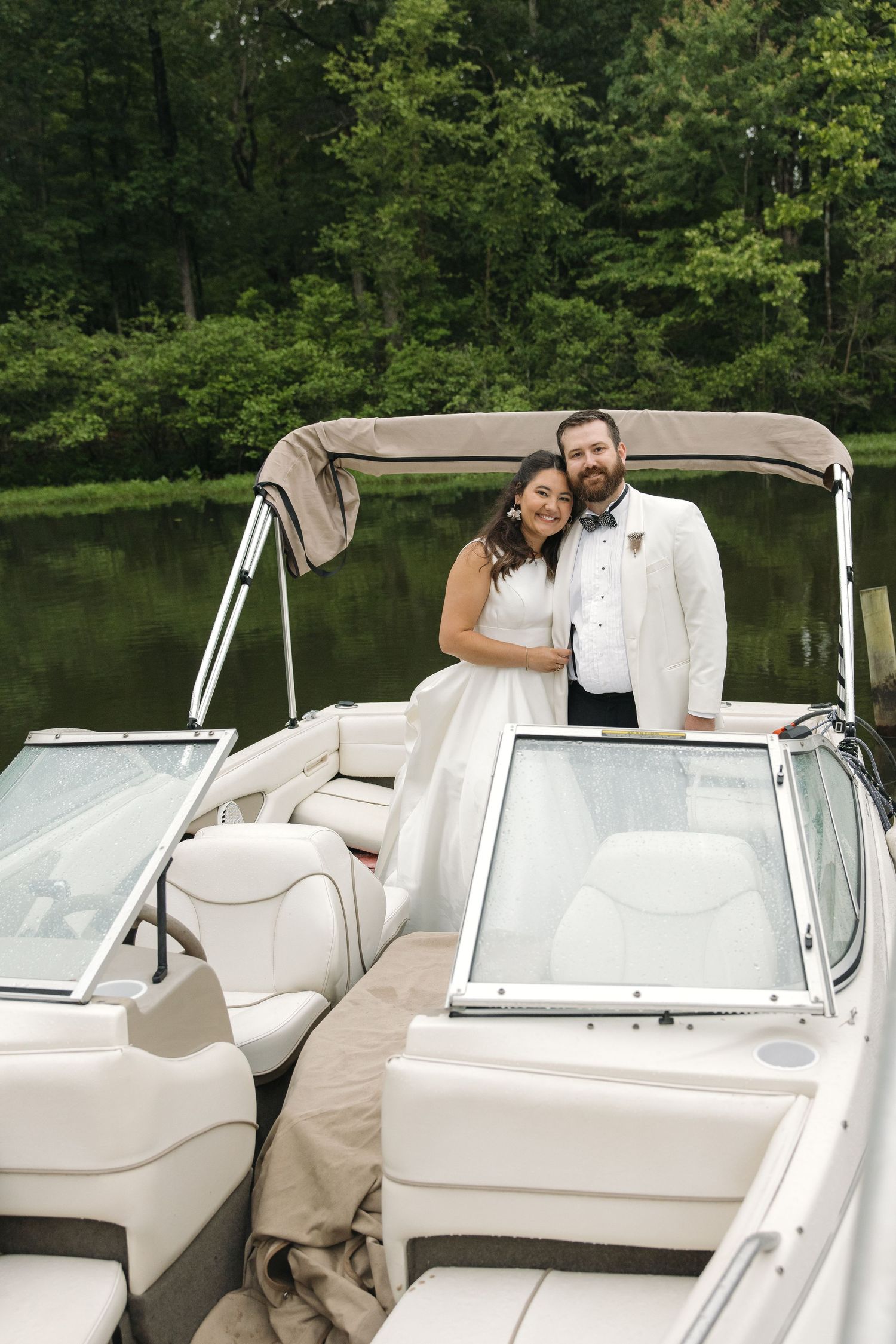 Bride and groom in white wedding attire smiling together on a luxury boat surrounded by lush green trees.