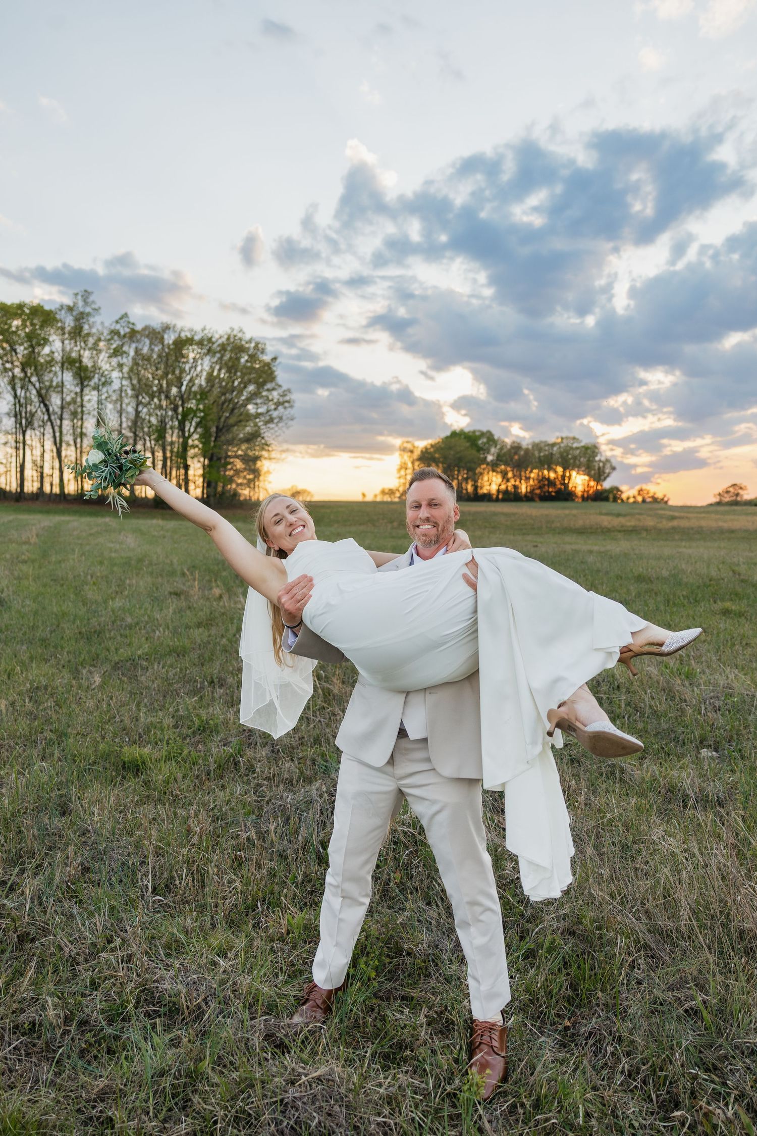 Groom carries laughing bride in his arms in a green field at sunset.