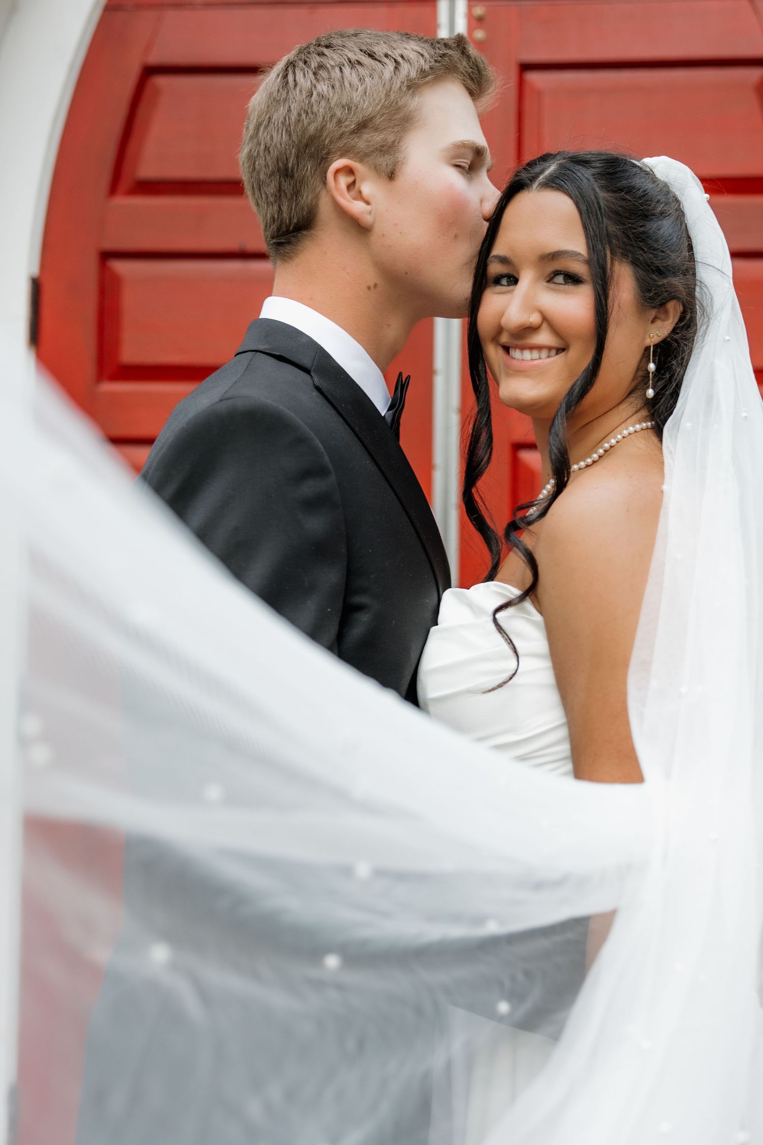 Groom in black tuxedo kissing smiling bride in white veil in front of a red door on their wedding day.