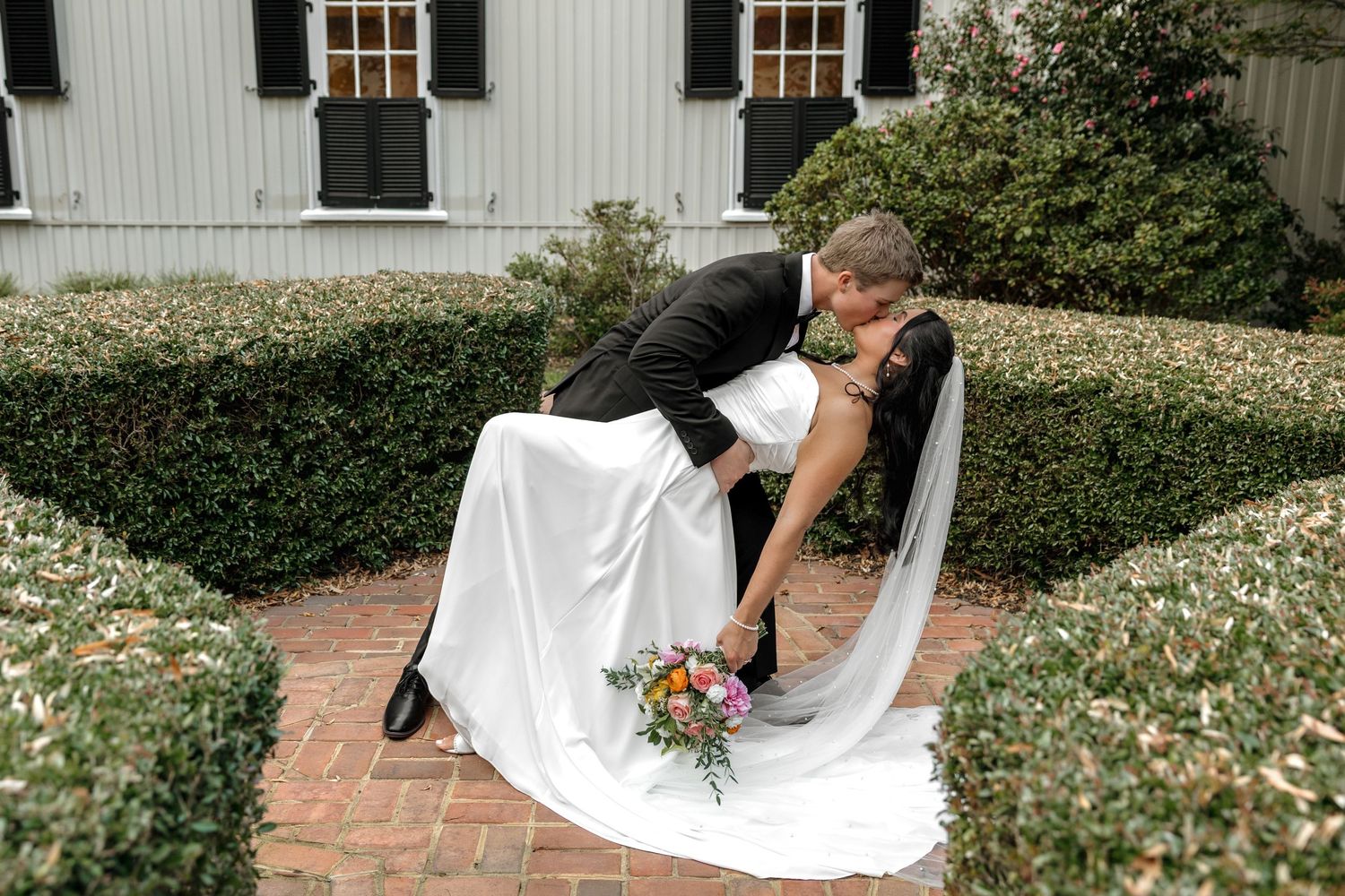 Groom dips bride for a romantic kiss in a garden courtyard with manicured hedges outside a white venue.