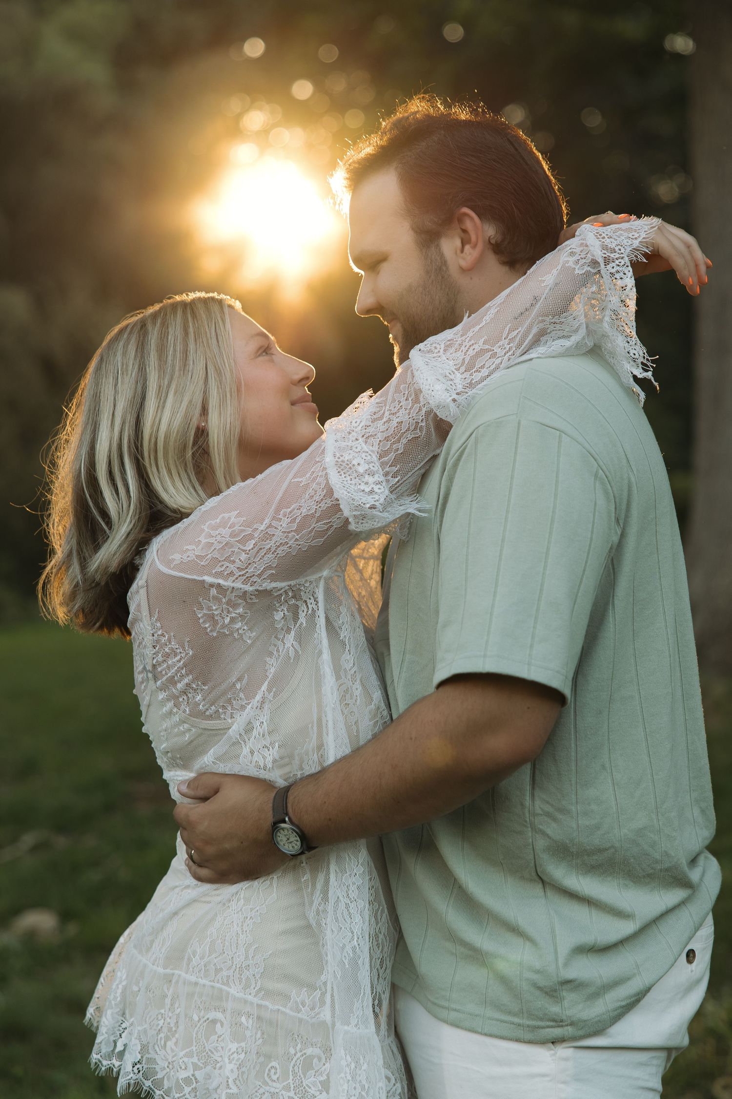 Couple embracing and smiling at each other during golden hour sunset in green park, woman in white lace dress.