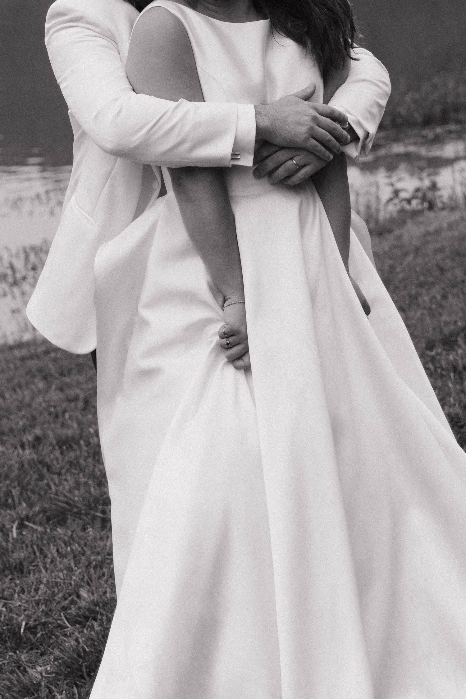 Black and white photo of groom embracing bride in elegant white wedding gown near a tranquil lake.