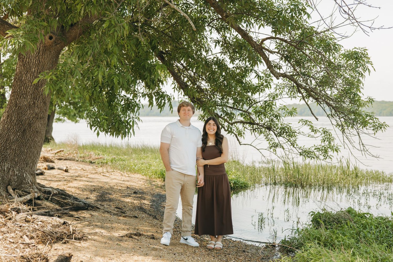 Couple posing together under a large tree beside a serene lake on a sunny day.