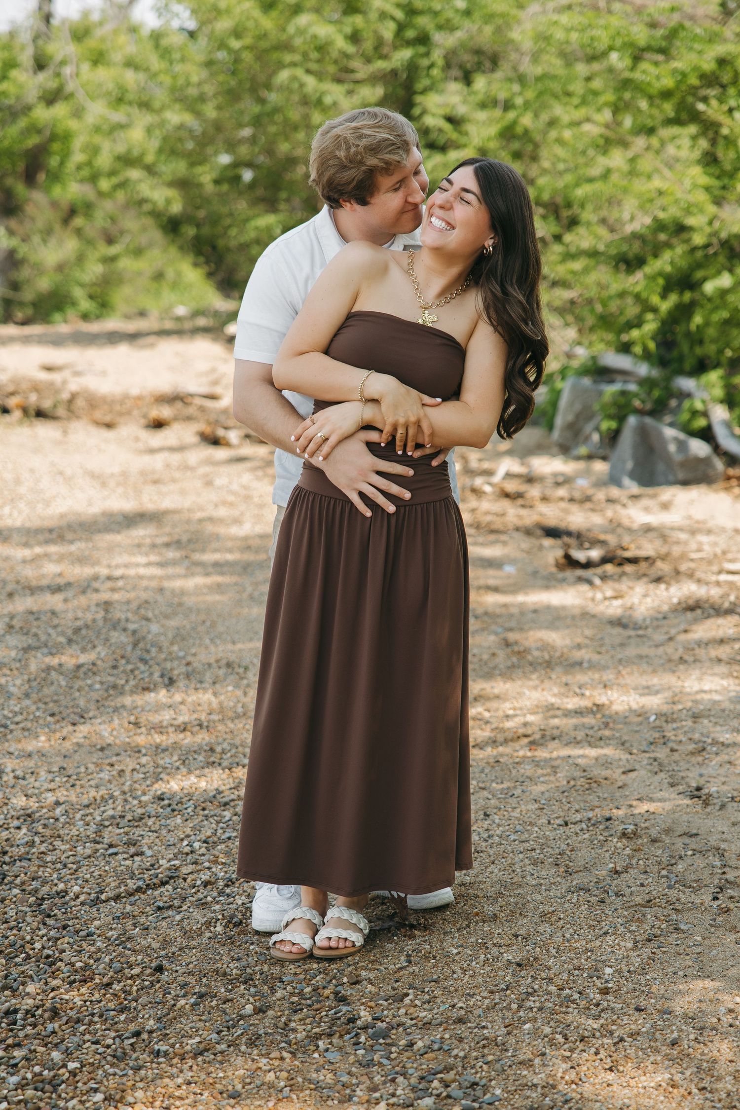 A couple embraces warmly outdoors, woman in brown strapless maxi dress laughing as man holds her from behind.