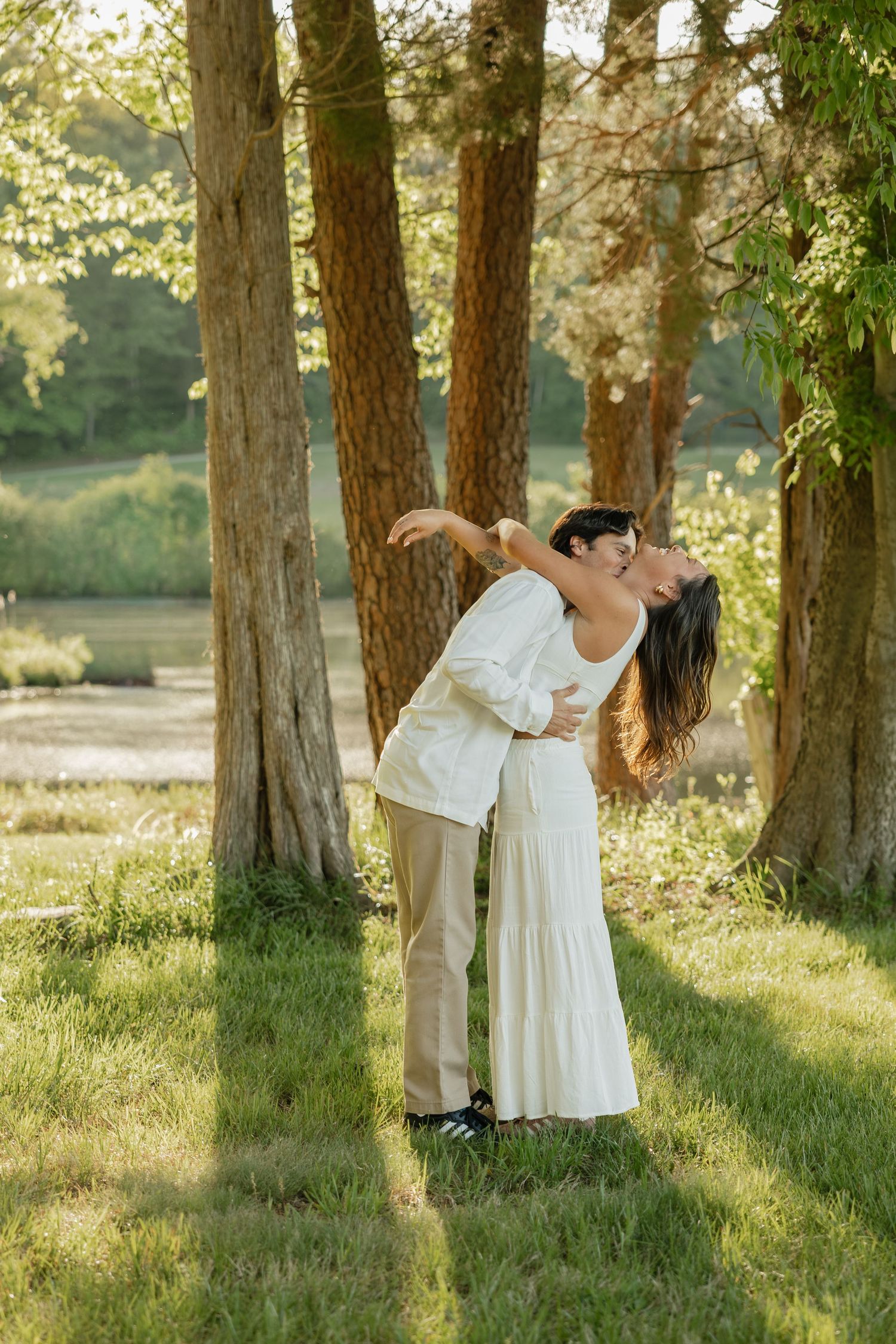 Couple sharing a romantic dip kiss among tall trees with golden sunlight filtering through the leaves.