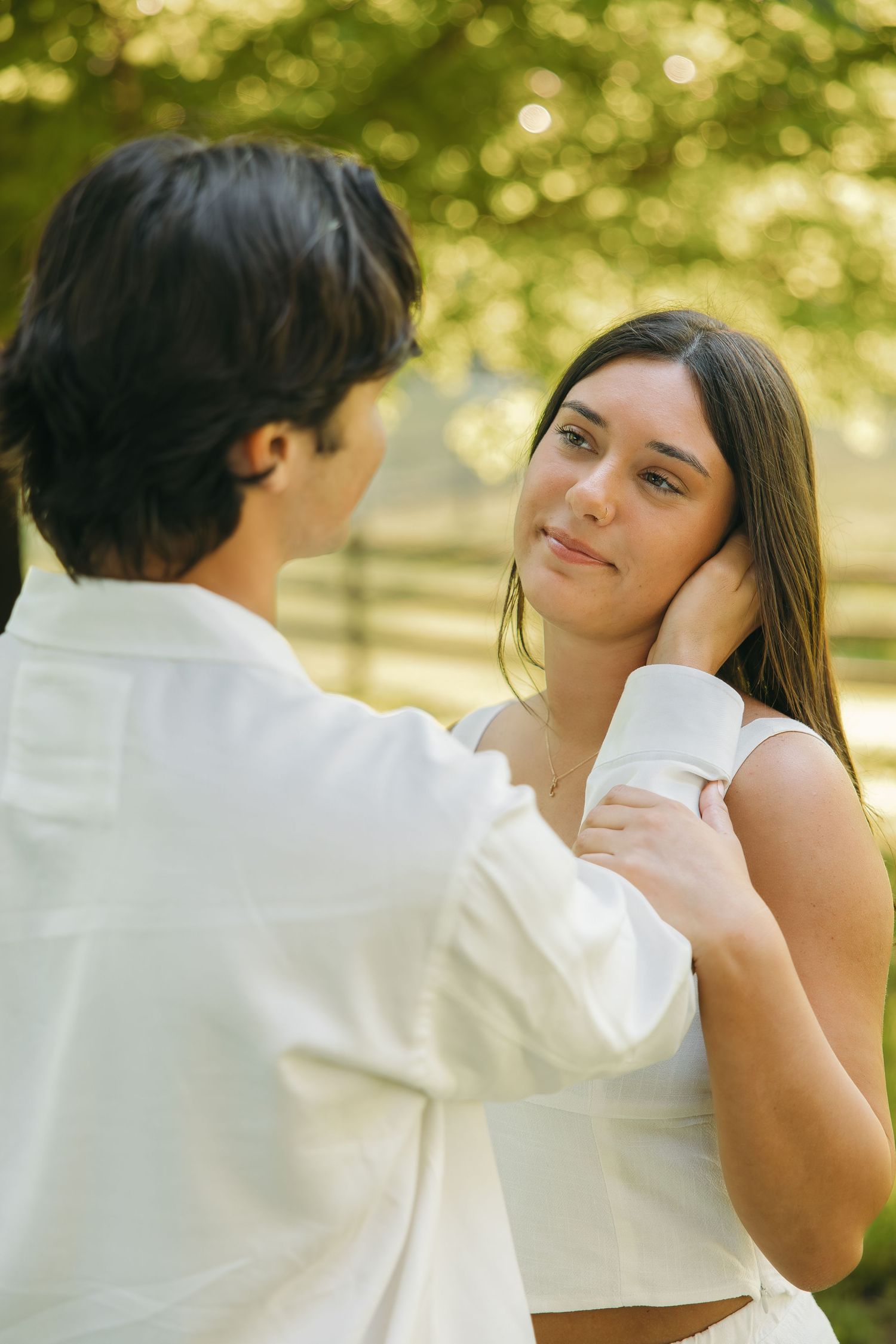 Man gently holding woman's face outdoors in warm sunlight, both dressed in white near a wooden fence.