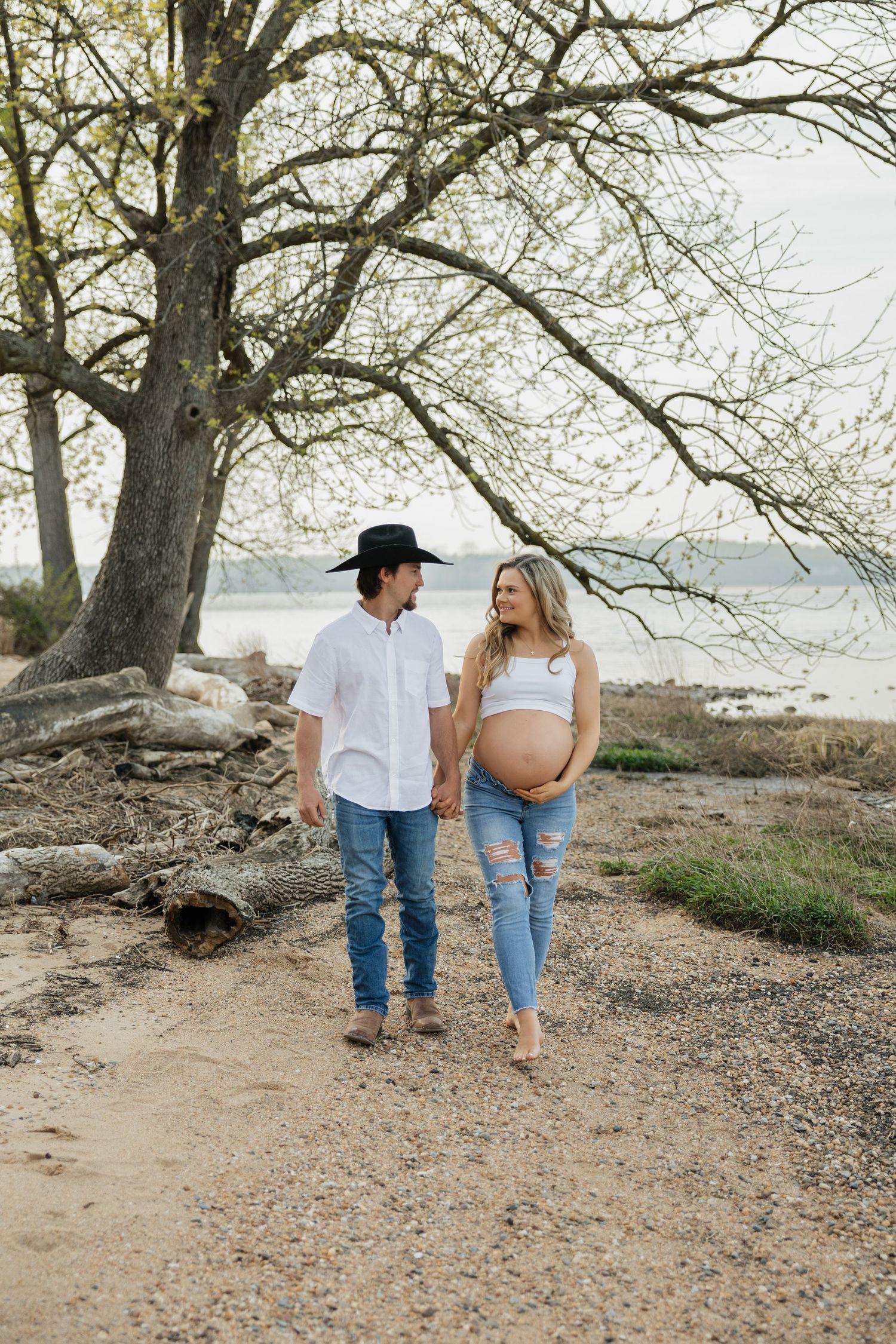 Pregnant couple holding hands walking along sandy path near water, man in cowboy hat and white shirt.