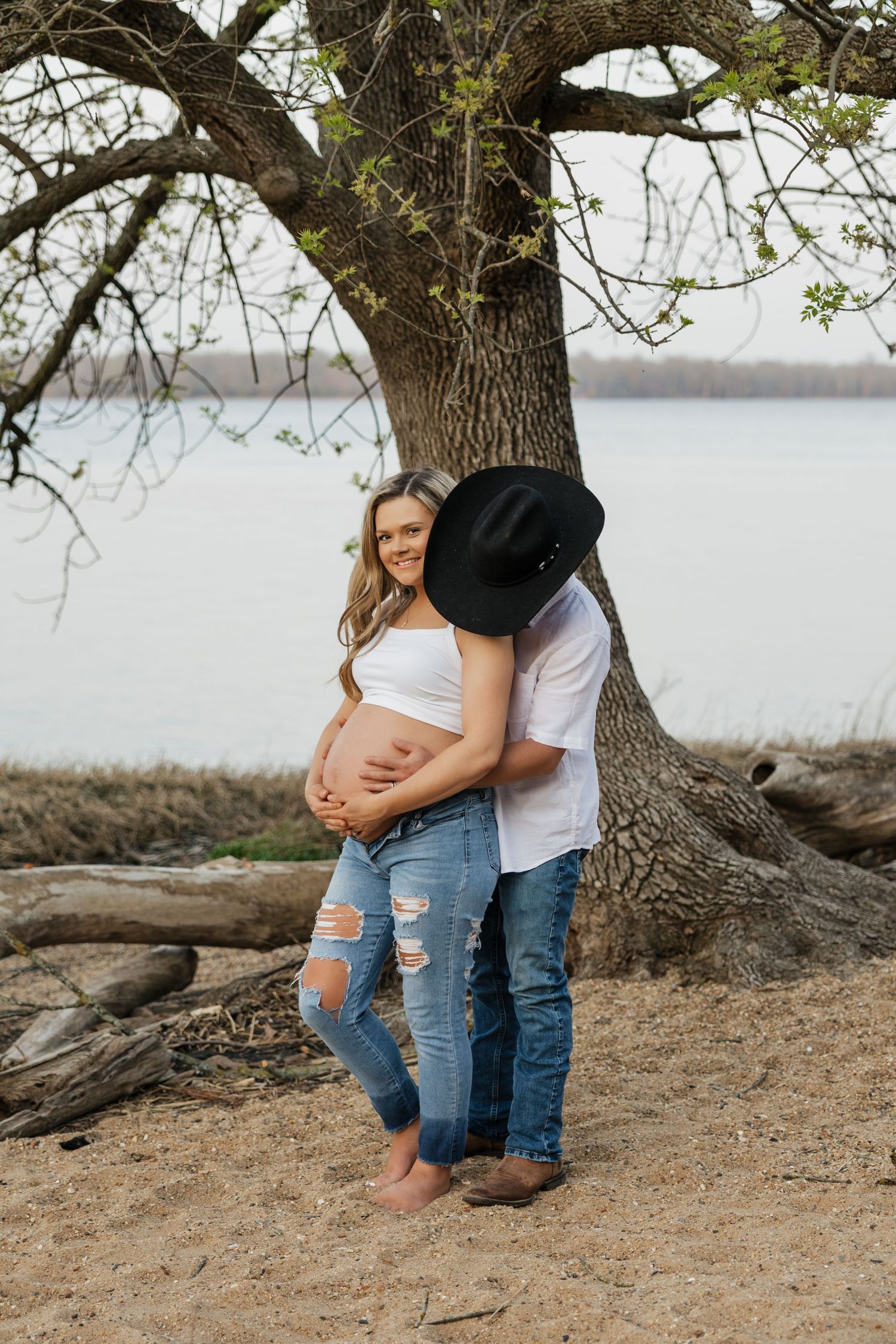 Couple embracing under tree by lake, pregnant woman in ripped jeans with partner in cowboy hat holding her belly.