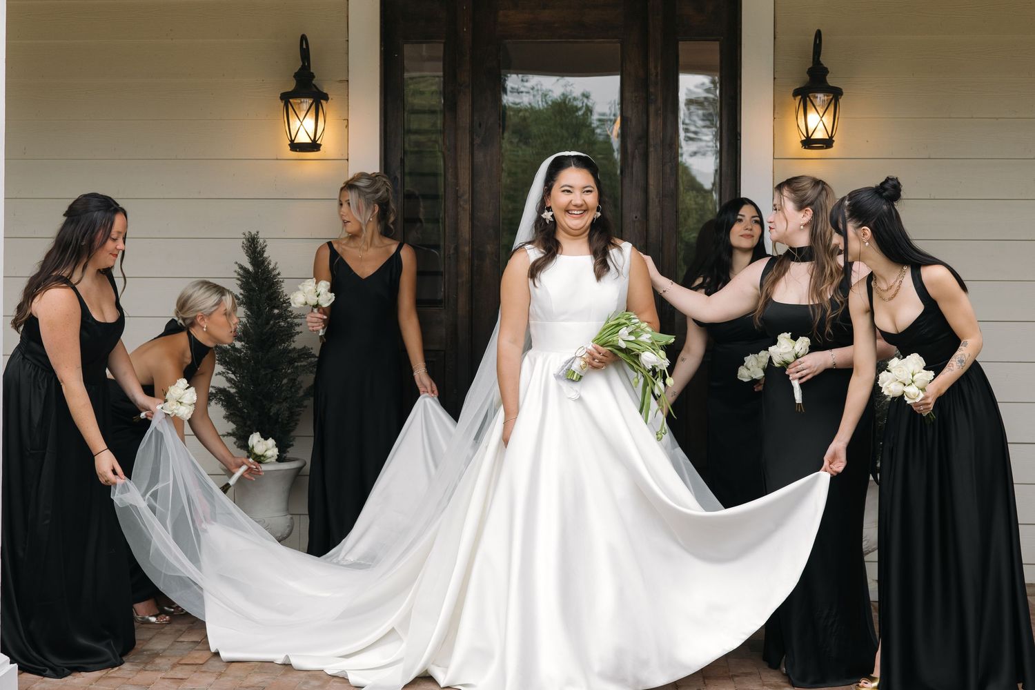 Smiling bride in white ball gown surrounded by bridesmaids in black dresses holding her veil and train outdoors.