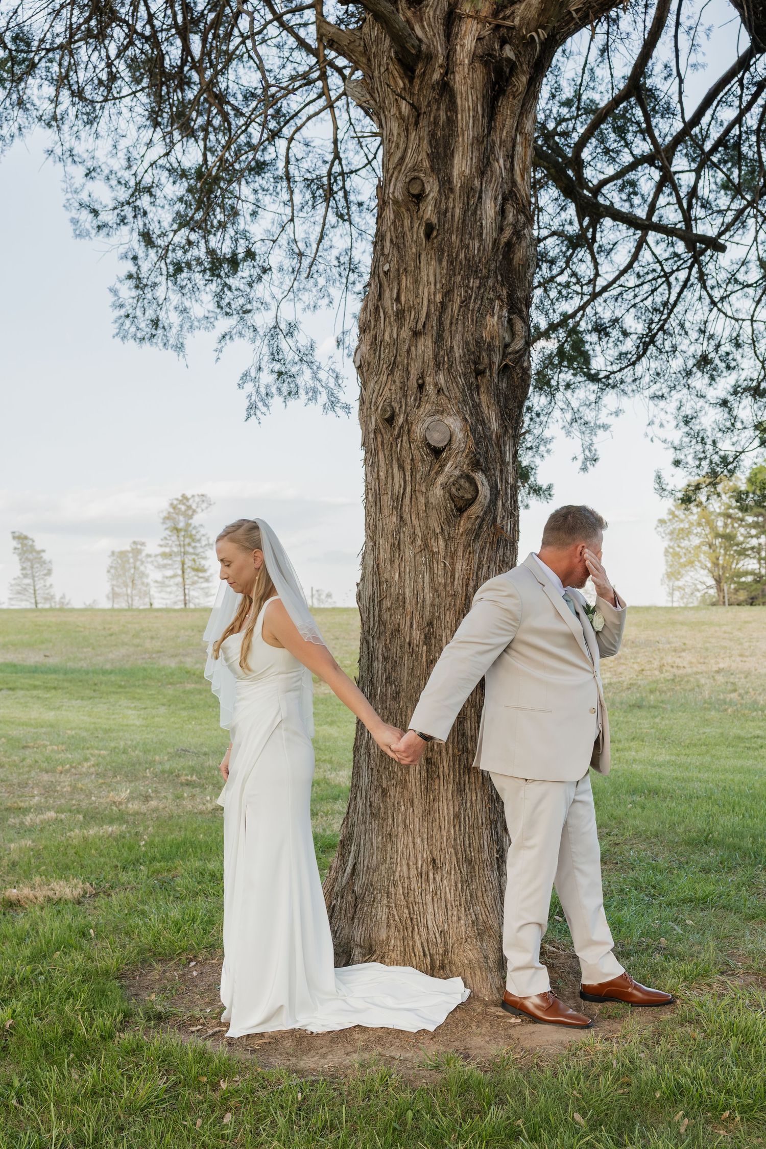 Bride and groom holding hands around a large tree trunk during wedding first look moment.