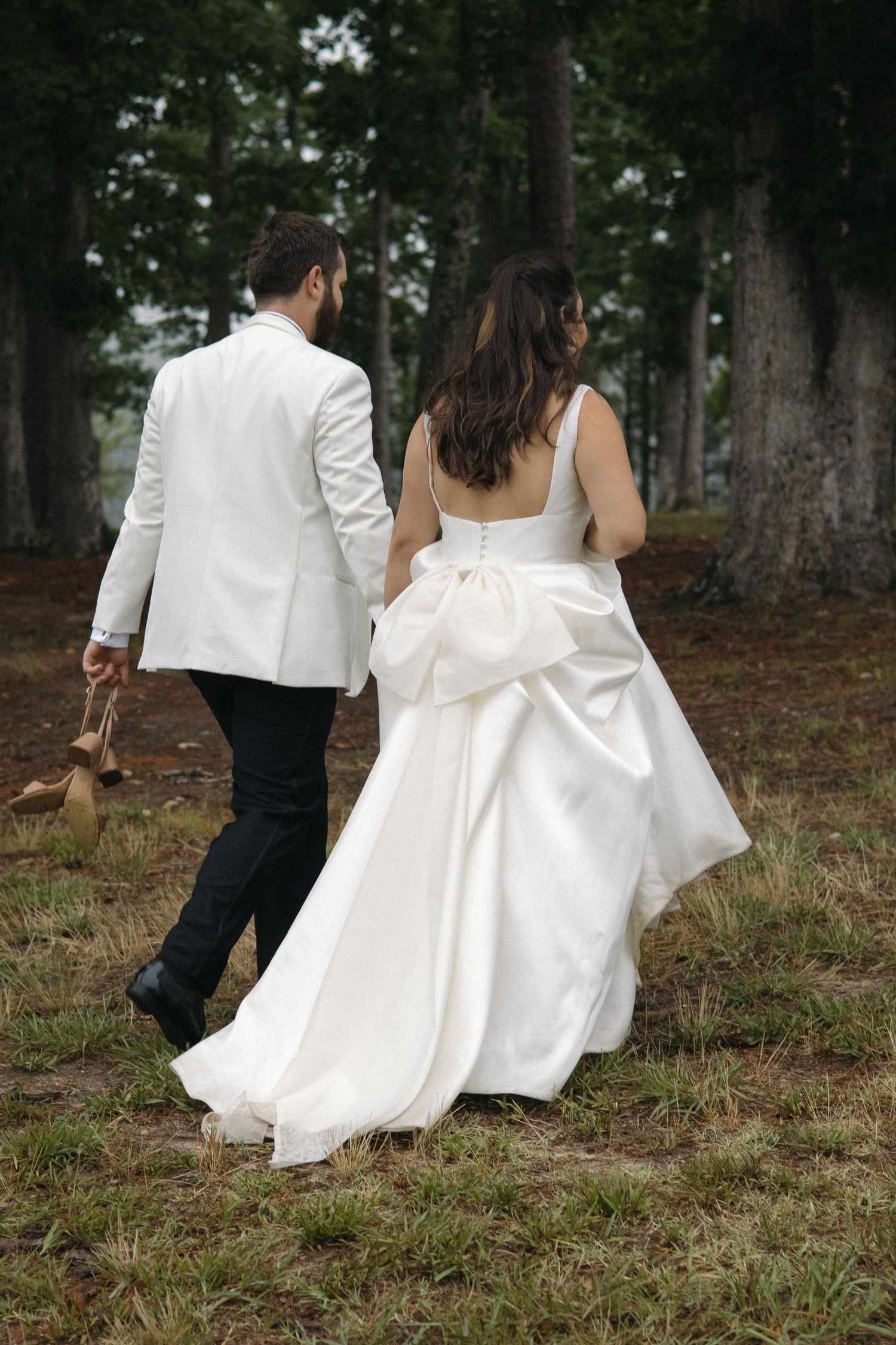 Bride and groom walk hand-in-hand toward tall trees, bride wearing gown with large bow detail.