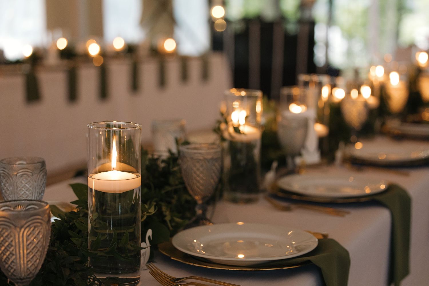 Romantic candlelit wedding table with greenery runner, glass votives, and sage green napkins.