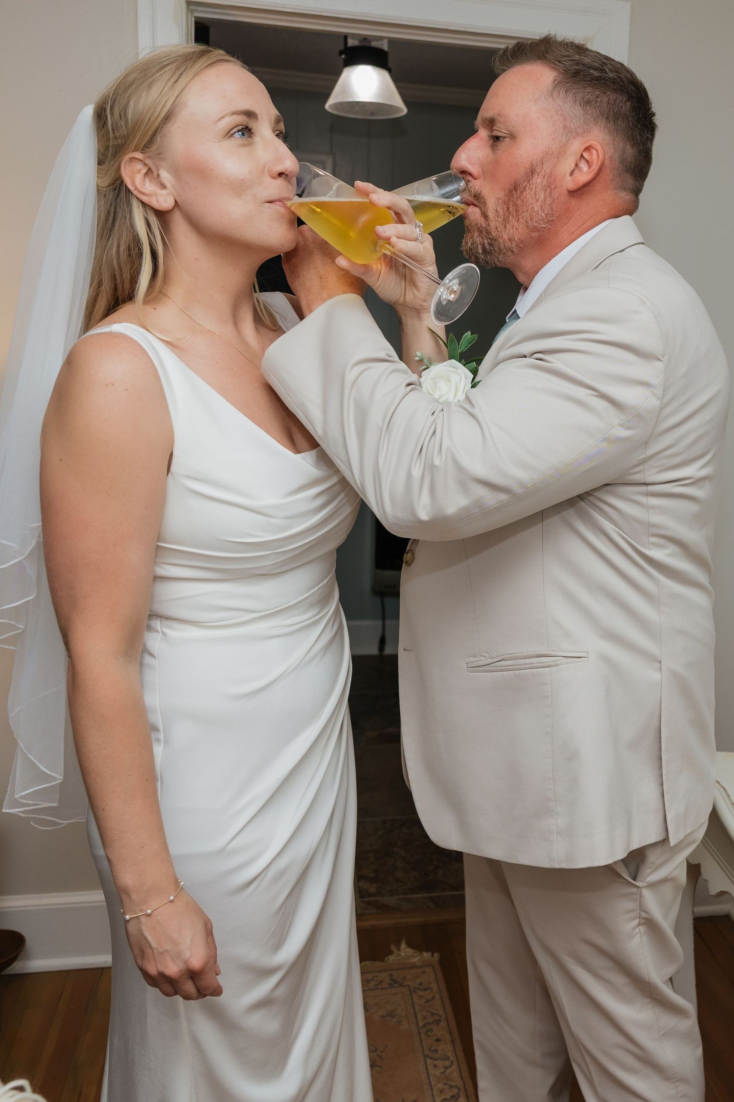 Bride and groom playfully share a champagne glass together, both drinking simultaneously in wedding attire.