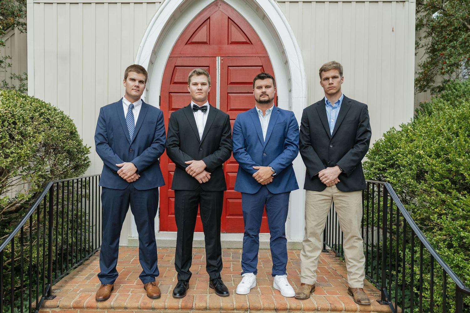 Groom in tuxedo poses with three groomsmen in suits before red church doors on wedding day.