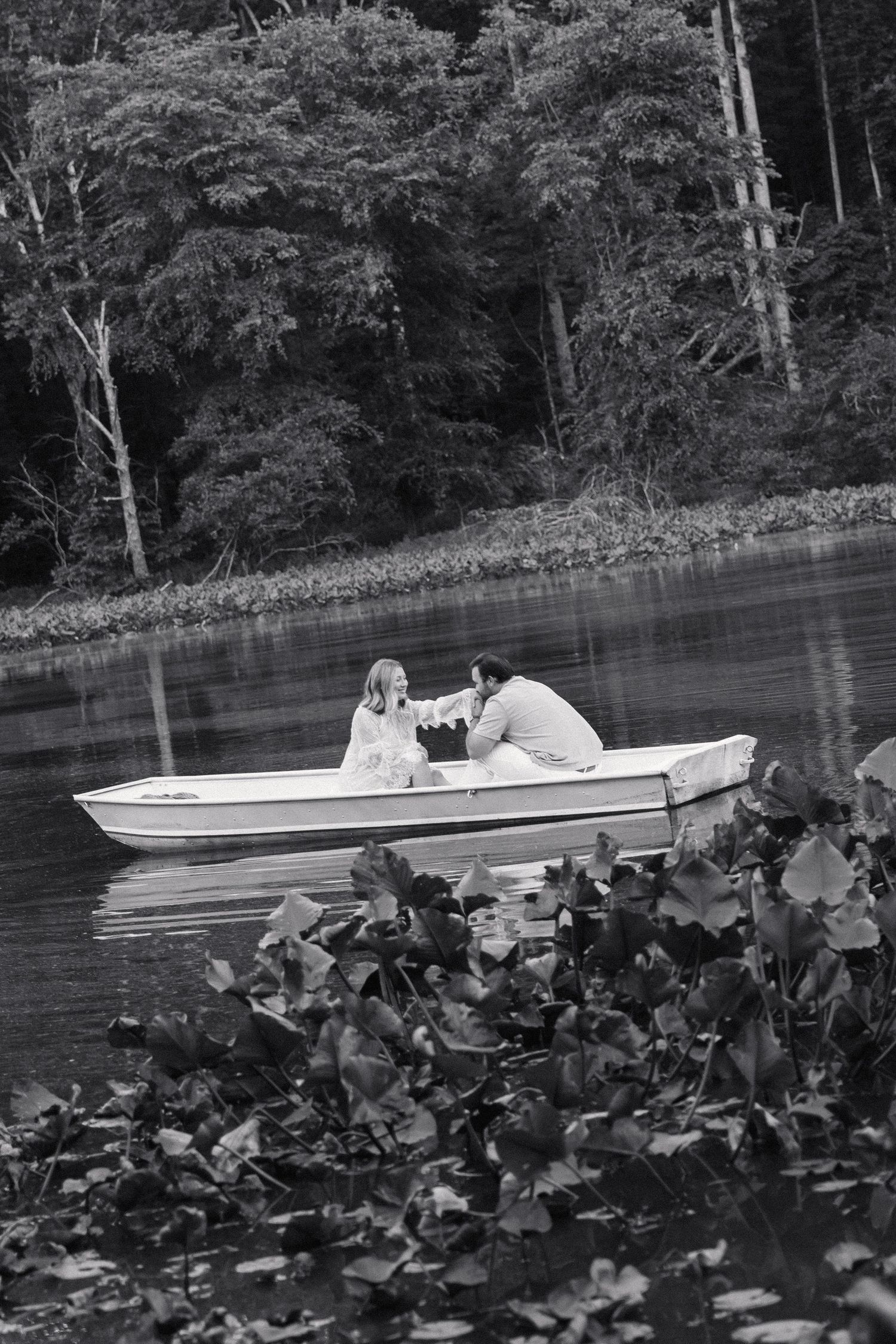 Black and white photo of a romantic couple sitting in a white rowboat on a calm lake surrounded by trees.