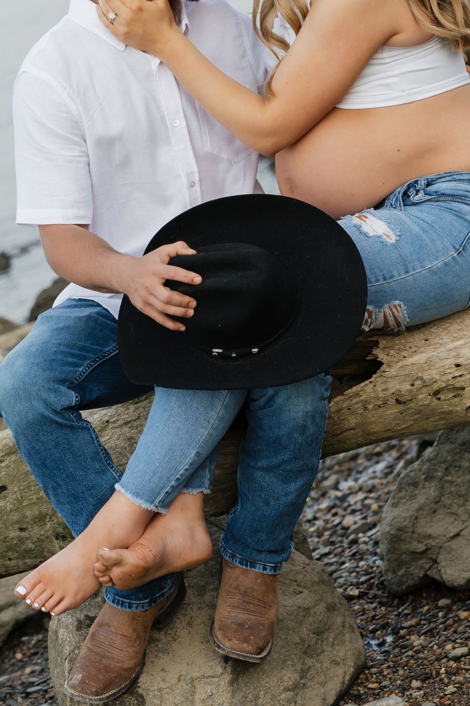 Close-up of couple sitting on driftwood, man holding black cowboy hat beside pregnant womans bare belly.