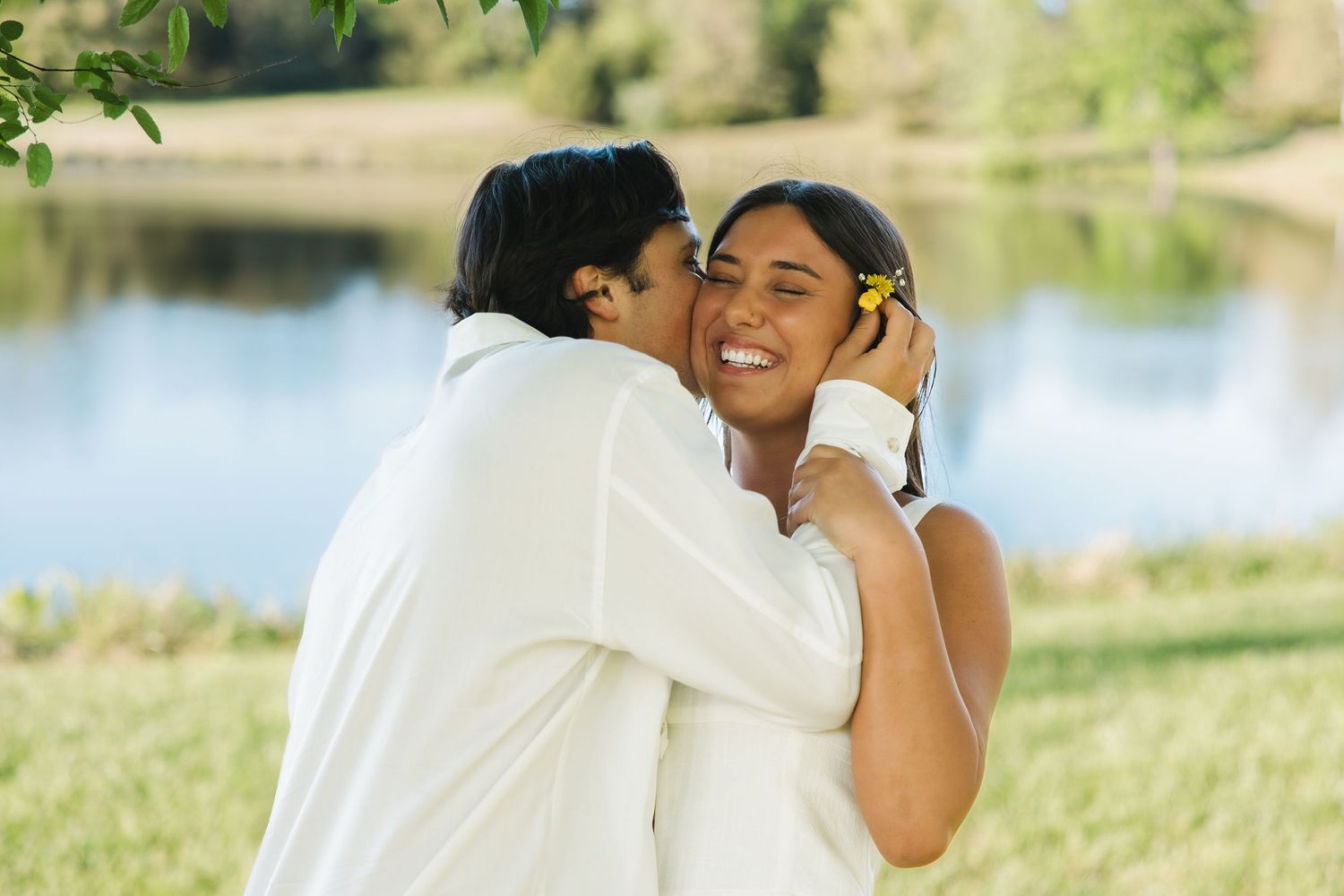 Man in white shirt kissing smiling woman with yellow flower in hair outdoors near a sunlit green lake.