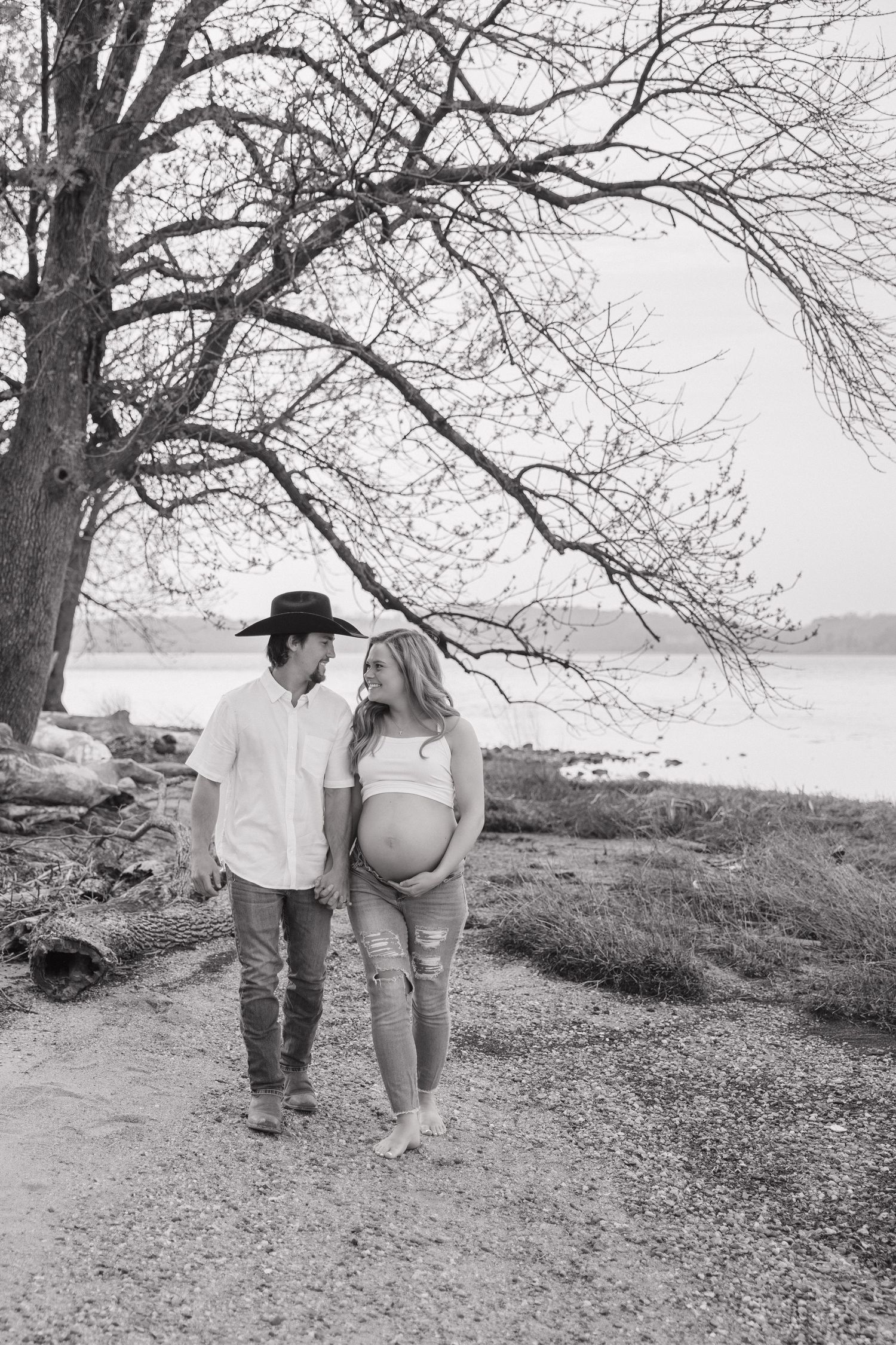 Black and white maternity photo of couple walking near lake, woman showing baby bump, man in cowboy hat.
