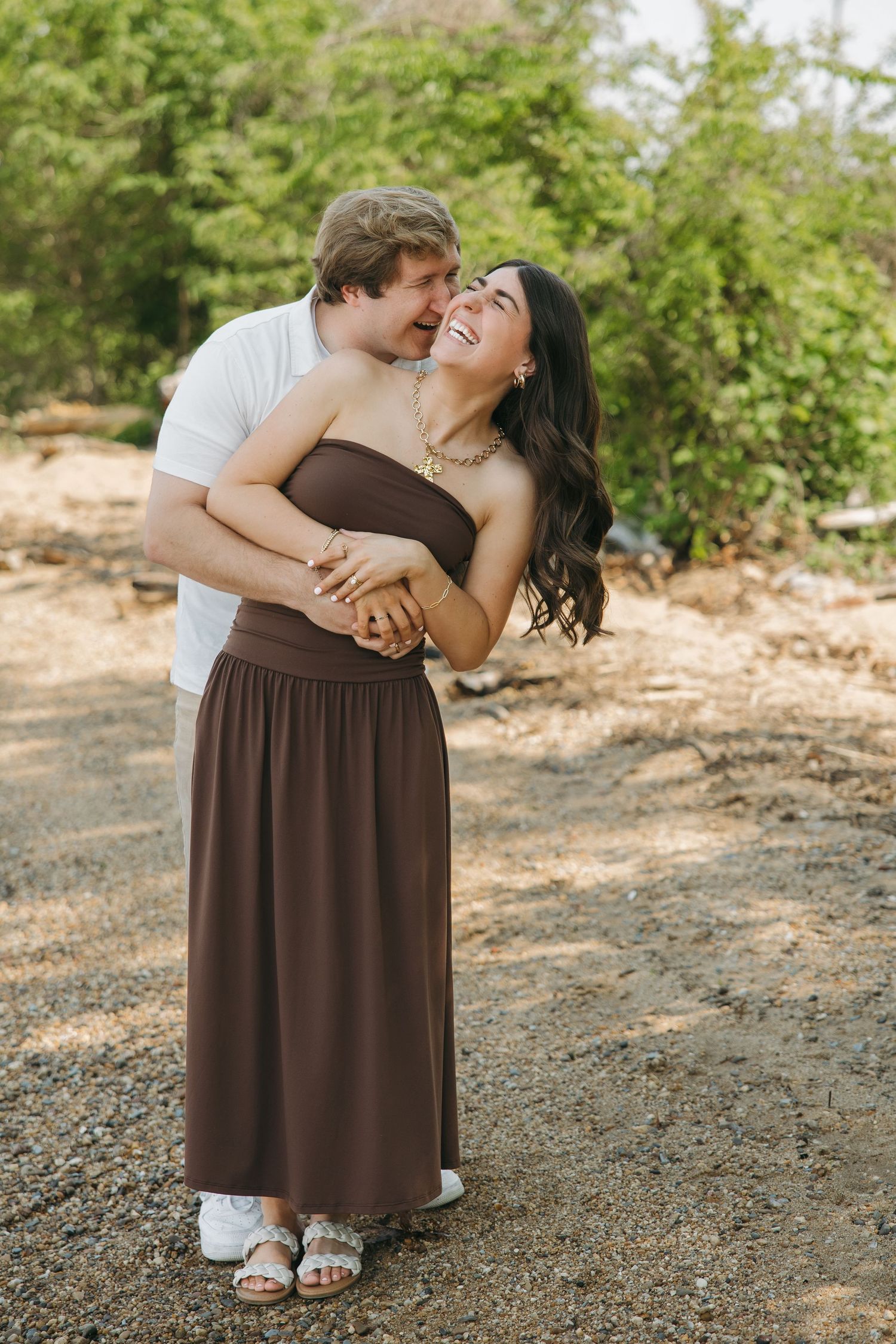 A joyful couple embraces on a gravel path, woman in brown strapless dress laughing as man holds her close.