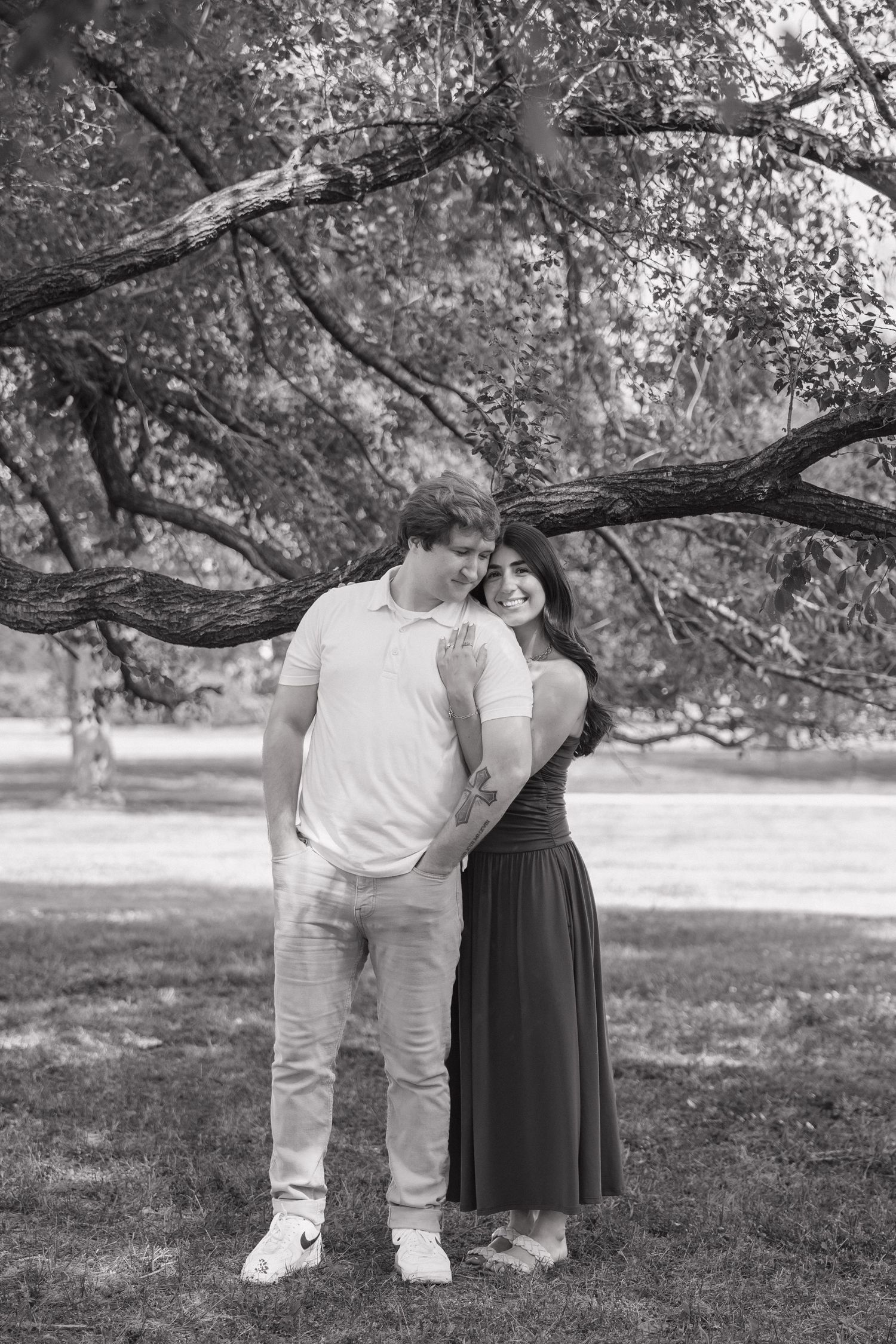 Black and white photo of a couple sharing a tender moment under a sprawling tree in a park setting.