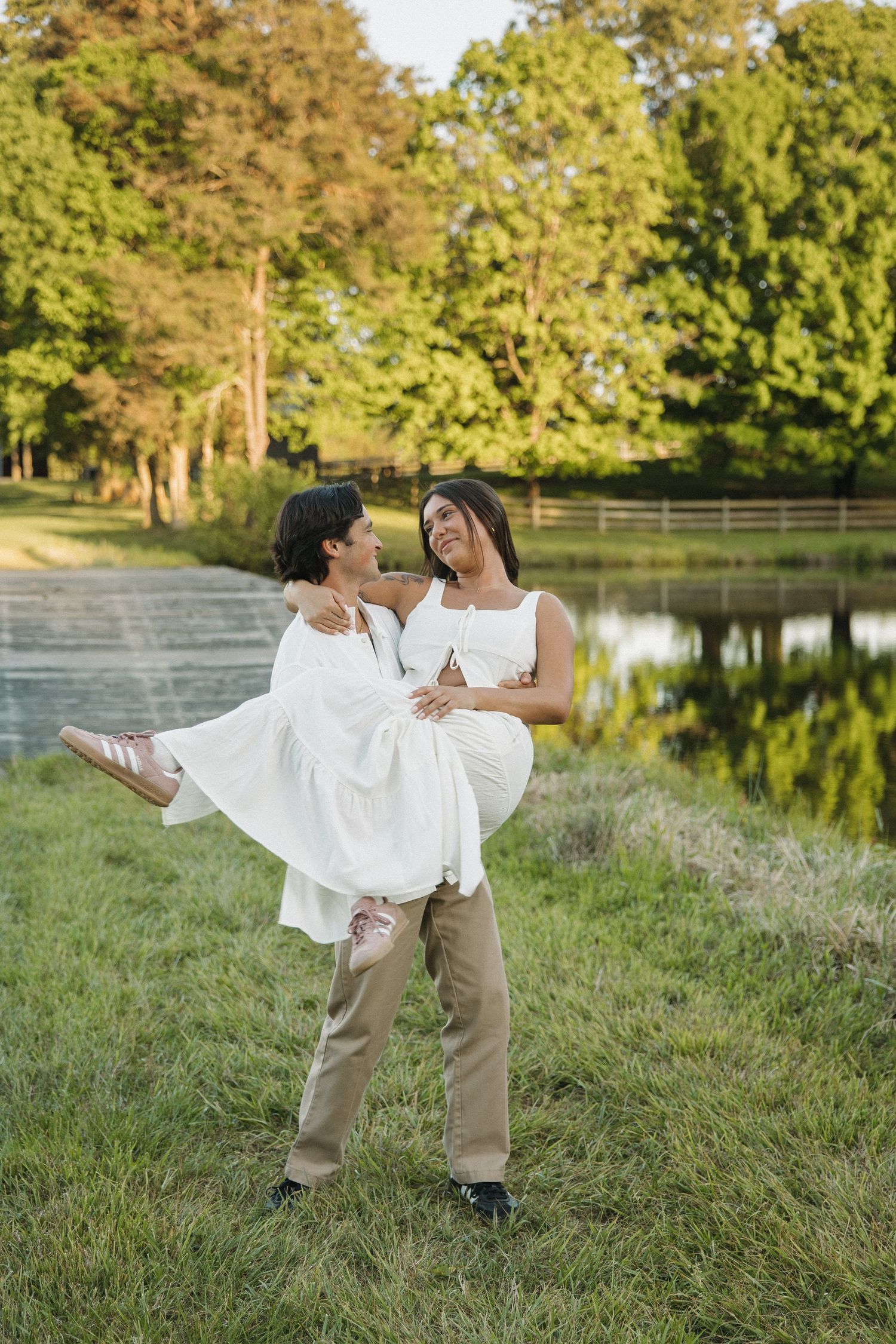 Man carrying woman in white dress by a serene pond surrounded by lush green trees on a sunny day.