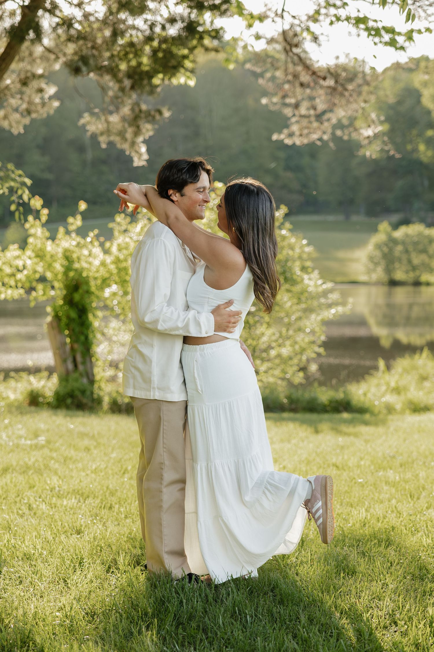 Happy couple embracing and smiling in a sunlit green meadow with a peaceful lake in the background.