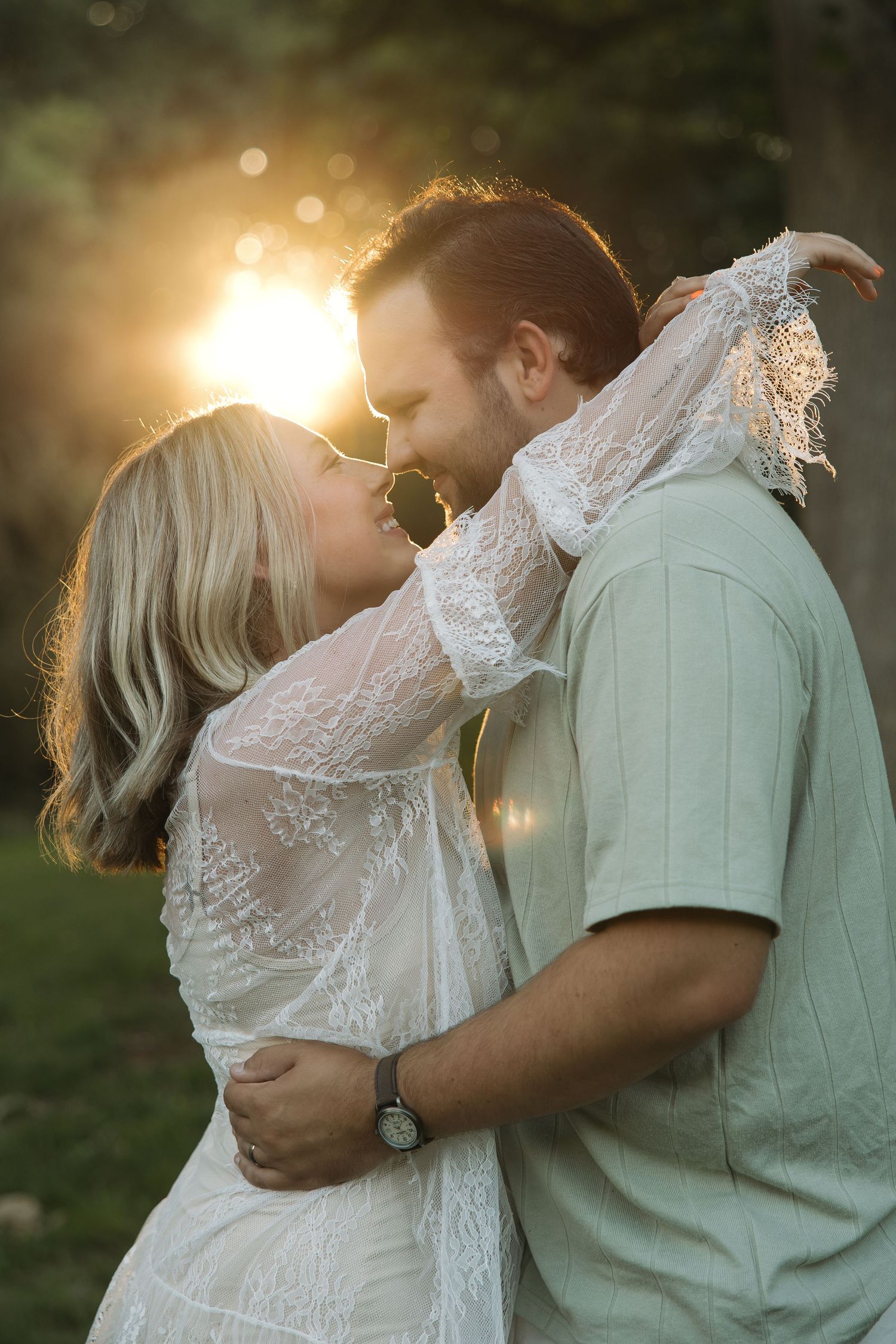 Couple sharing a tender kiss backlit by brilliant golden sunset, woman in white lace dress embracing man.