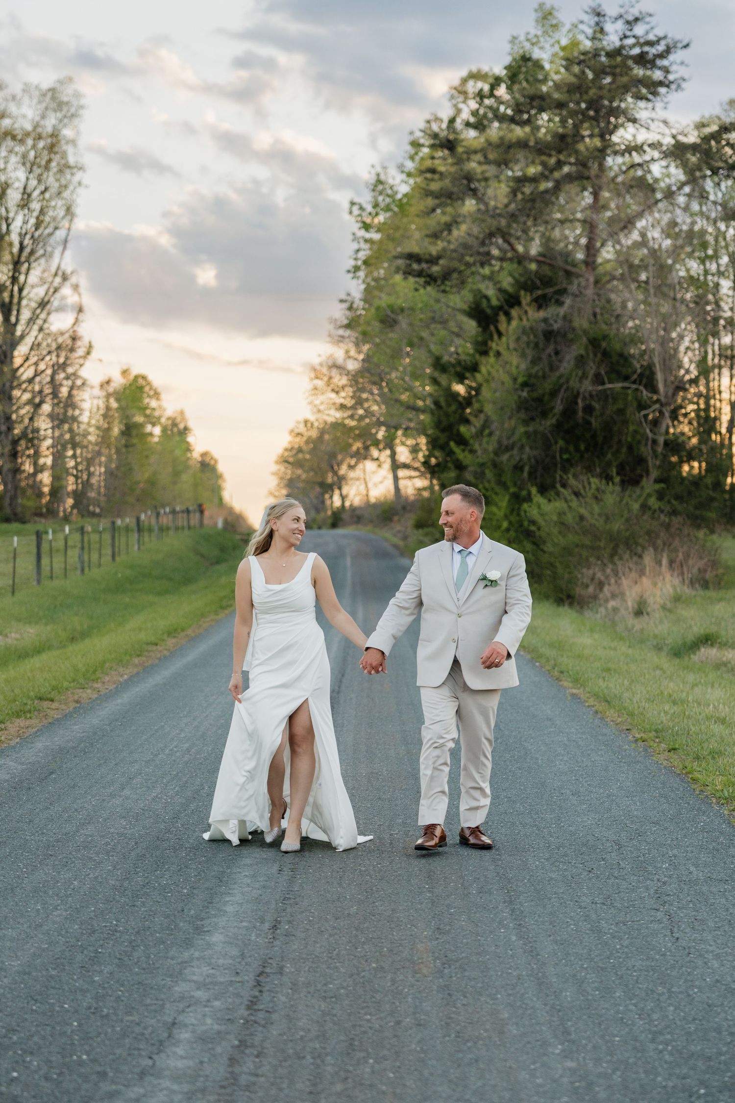 Bride and groom smile while walking hand in hand on a rural road at dusk.