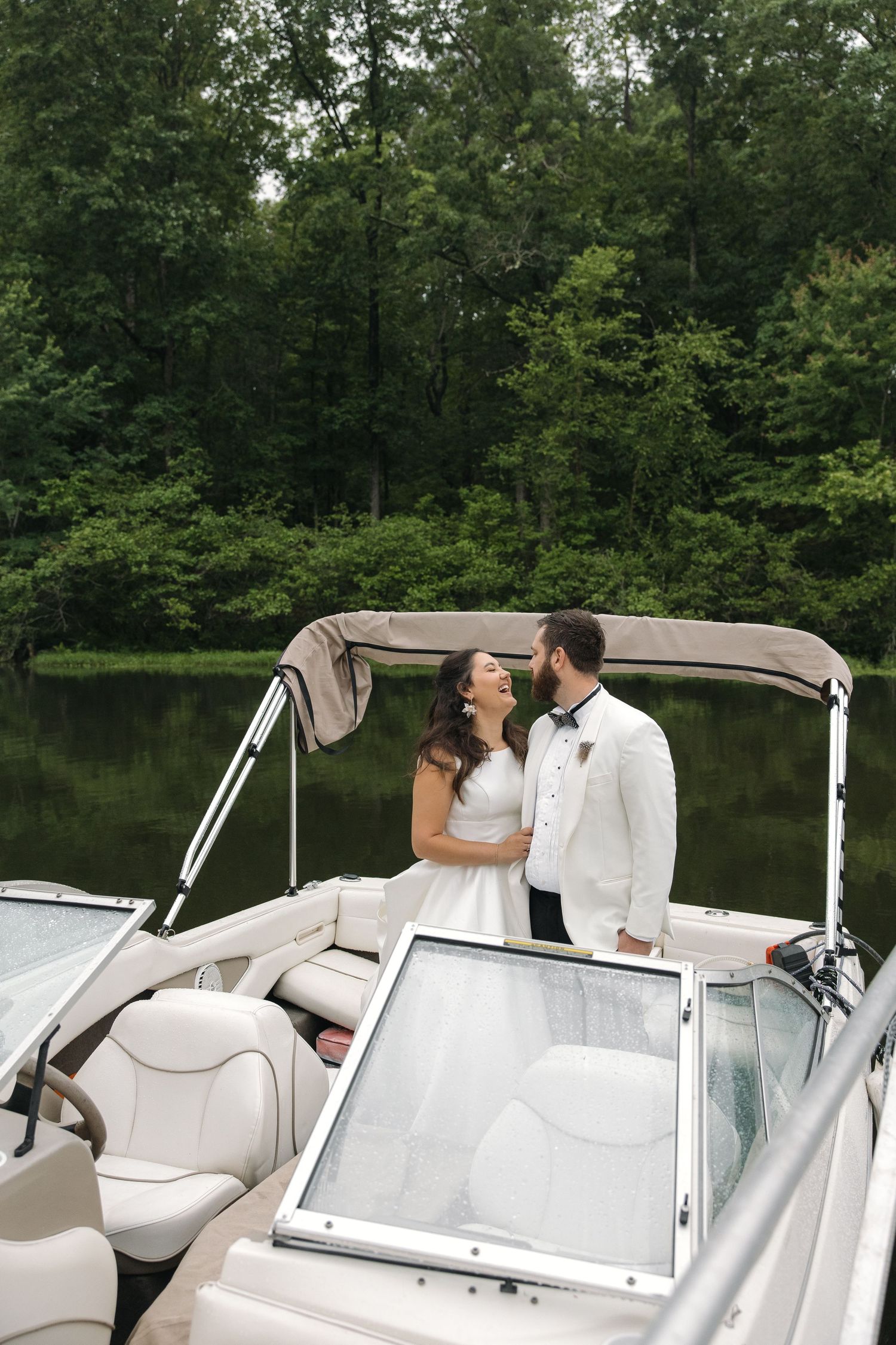 Bride in white gown and groom in white tuxedo laughing together on a luxury boat by a forest lake.