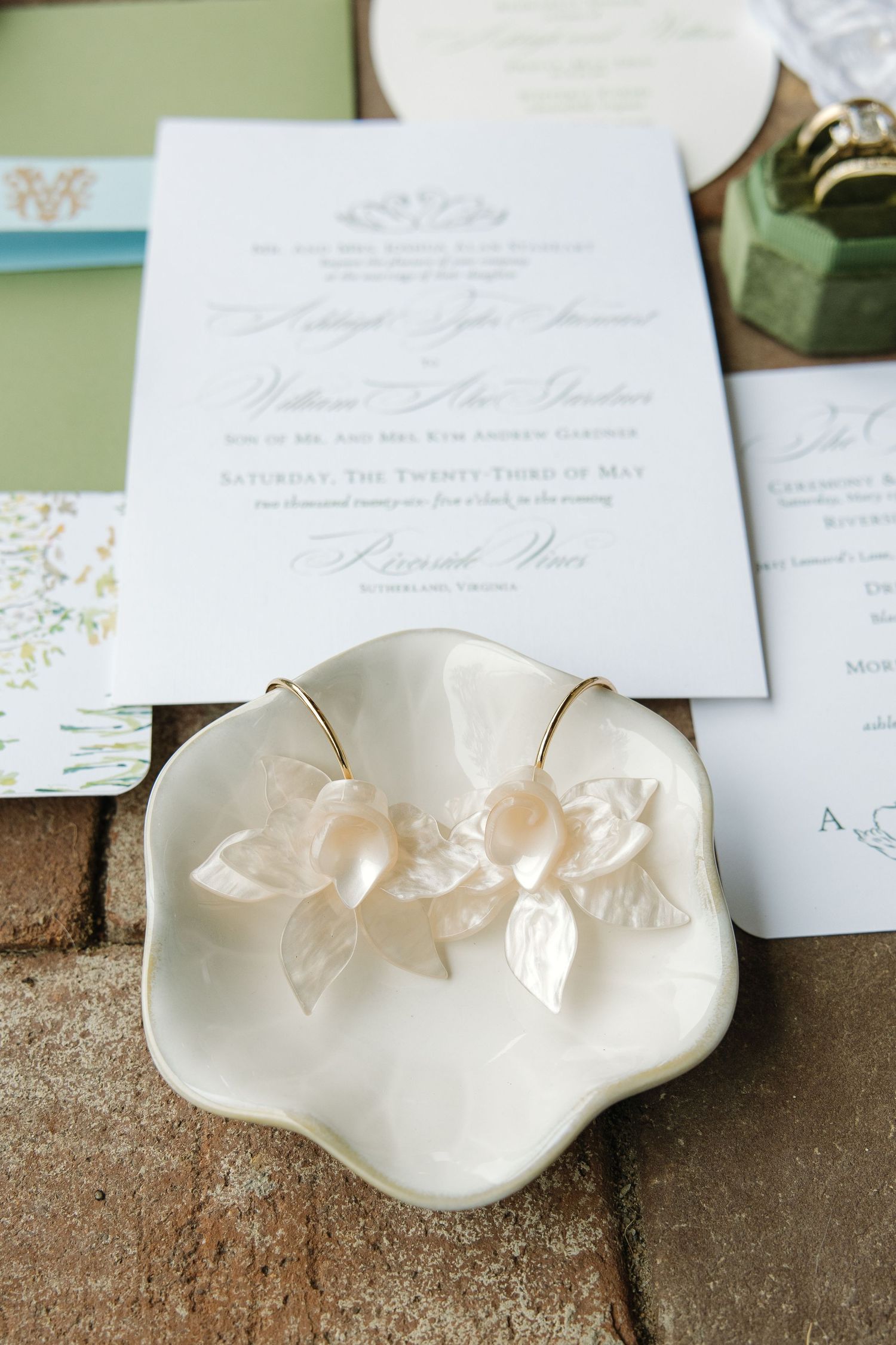 Delicate floral pearl earrings in white ceramic dish displayed alongside wedding stationery on brick surface.