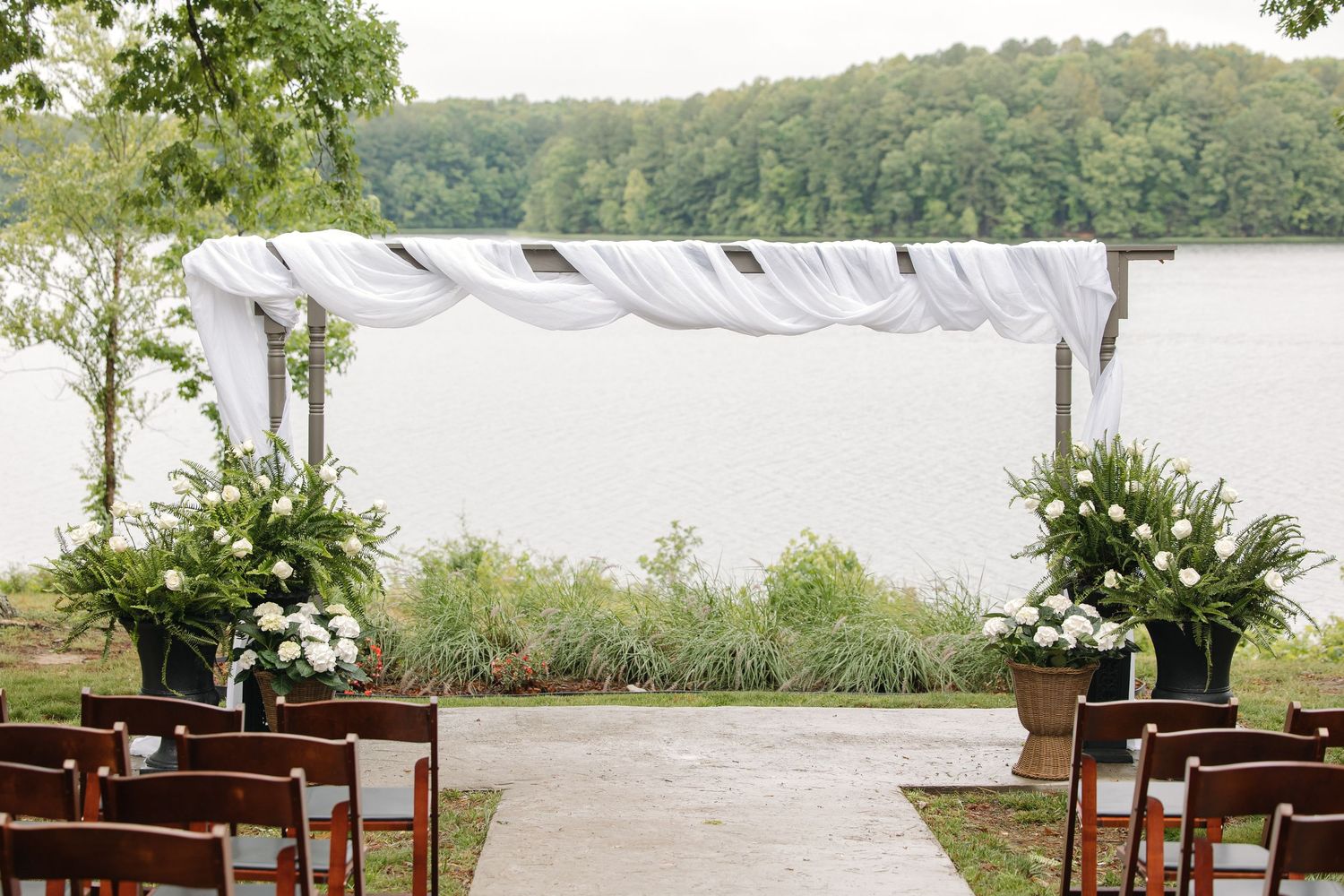 Elegant outdoor wedding ceremony setup with white draped arch and floral arrangements by a lake.