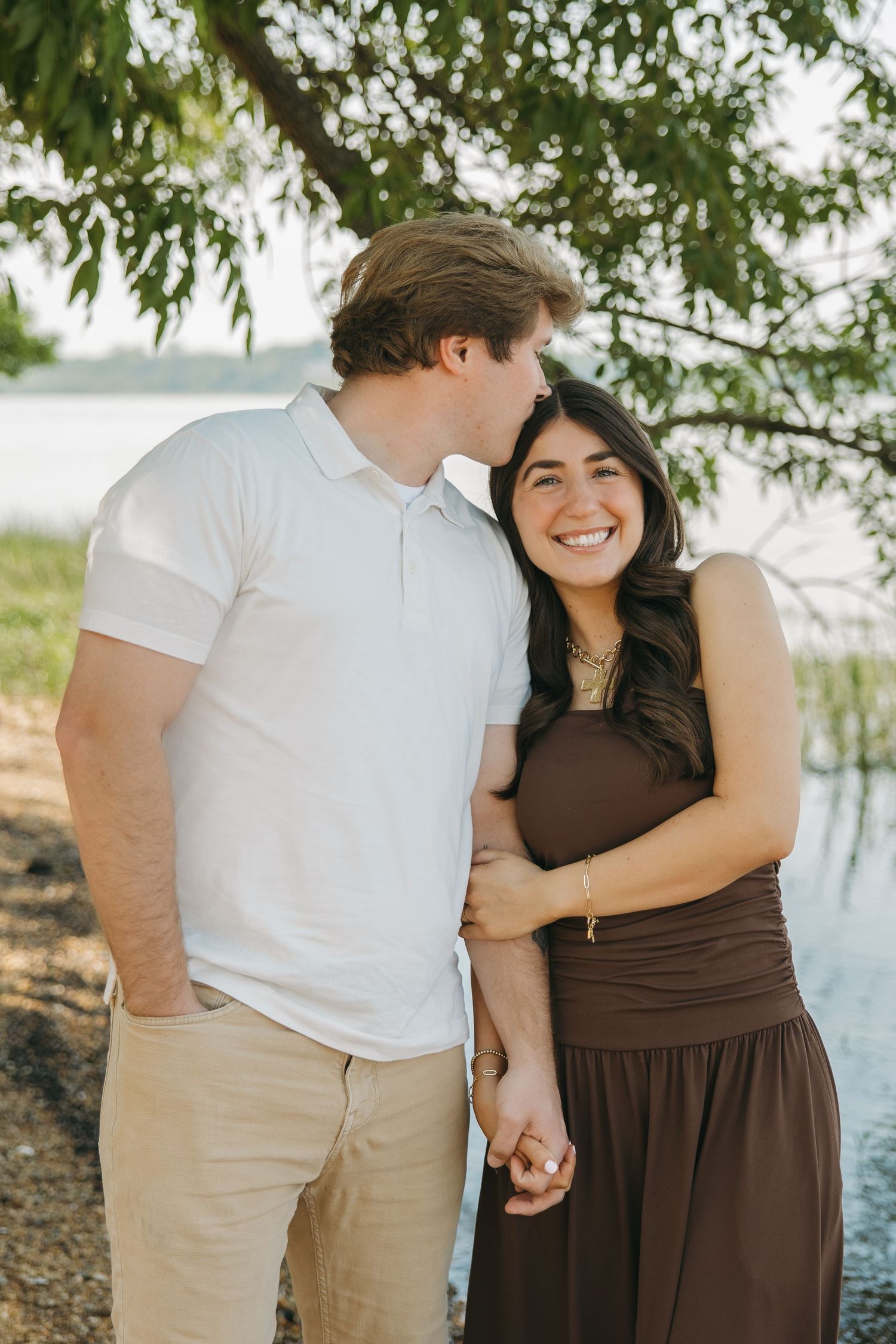 A man kisses a laughing woman on the cheek while holding hands near a sunlit lake under a tree.