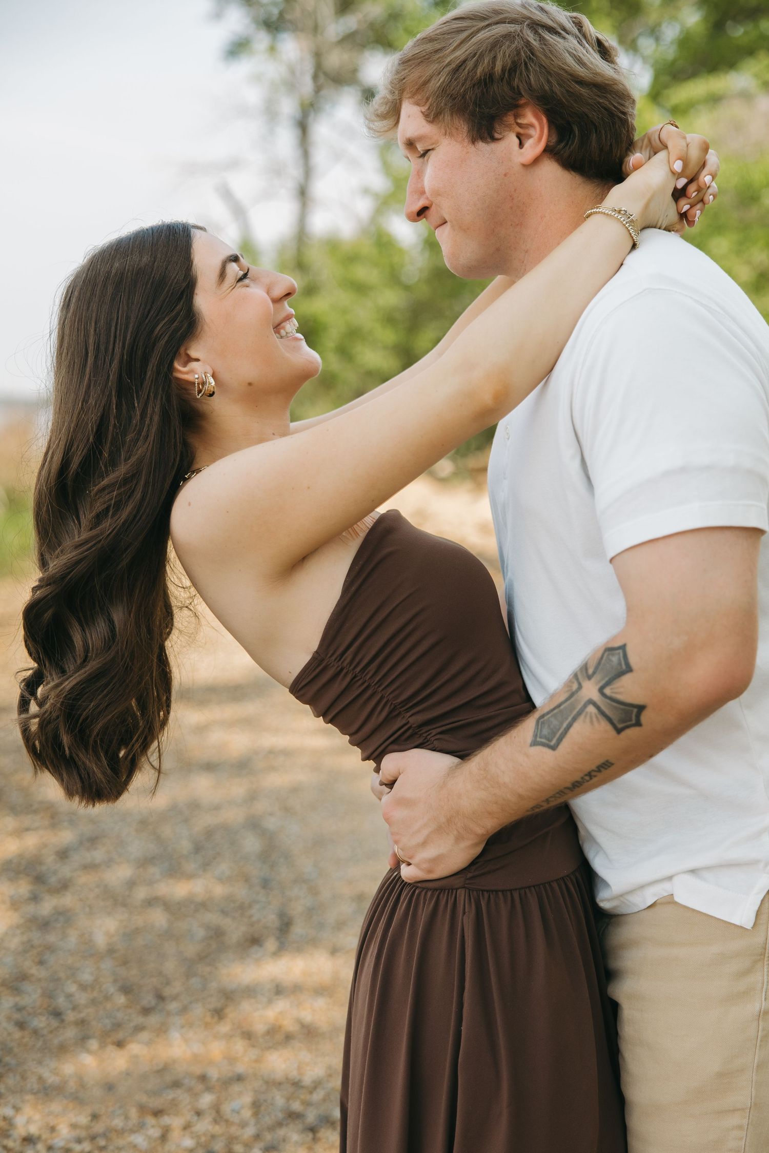 A smiling couple gazes at each other, woman in brown dress embracing tattooed man in white shirt outdoors.