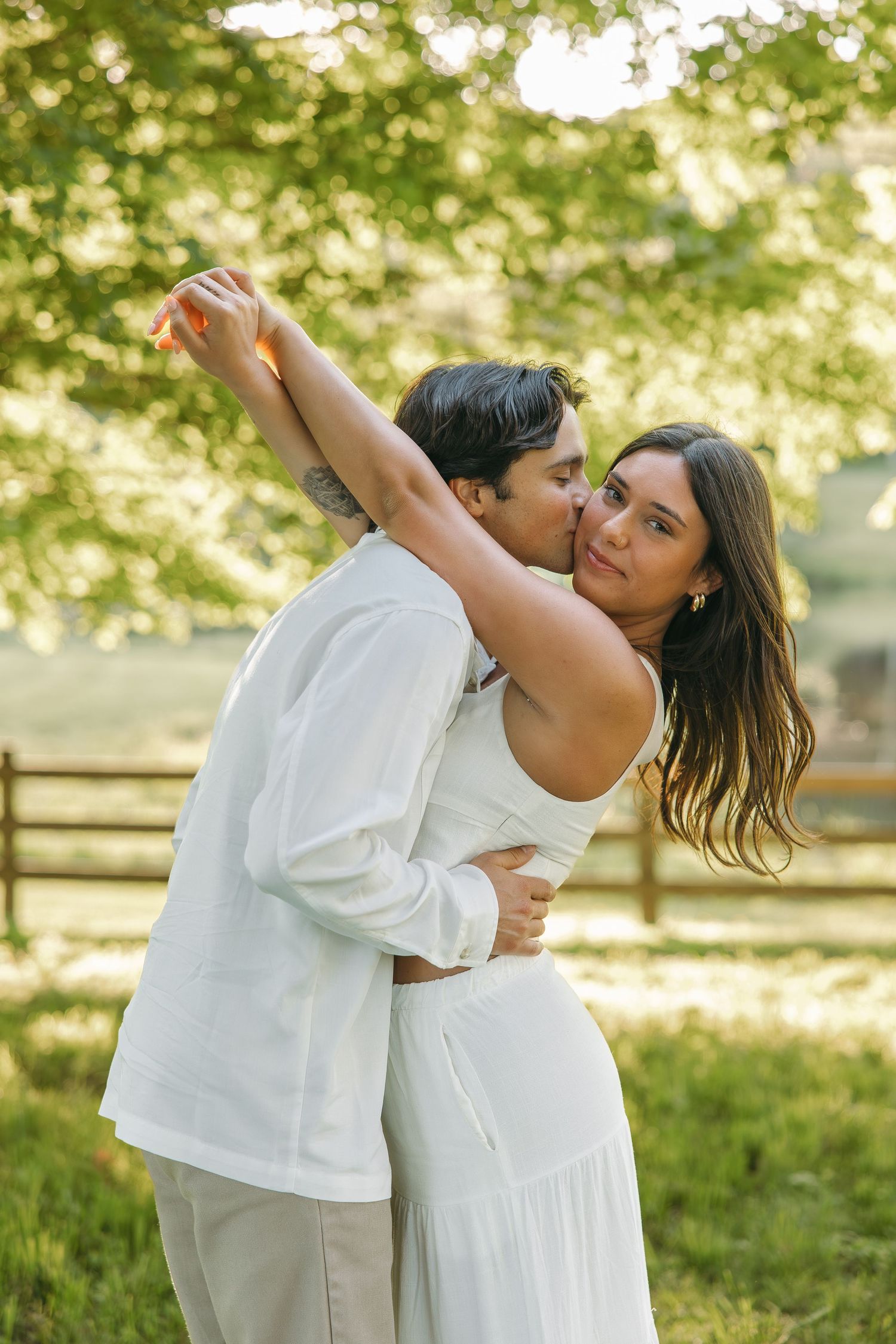 Couple sharing tender moment outdoors, man kissing woman's cheek as she raises her arm, green trees in background.