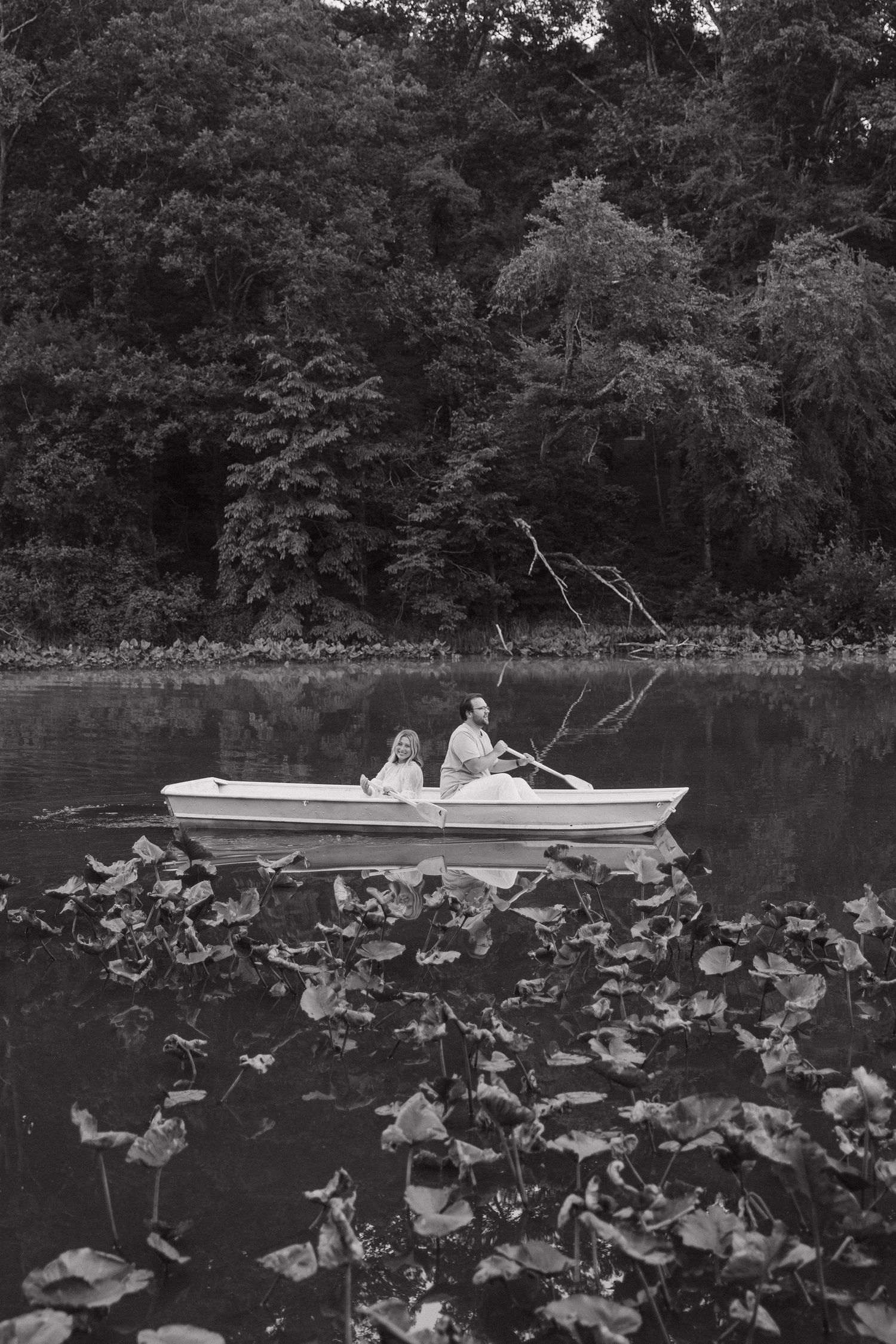 Black and white image of a couple in a white rowboat on a peaceful lake surrounded by trees and lily pads.