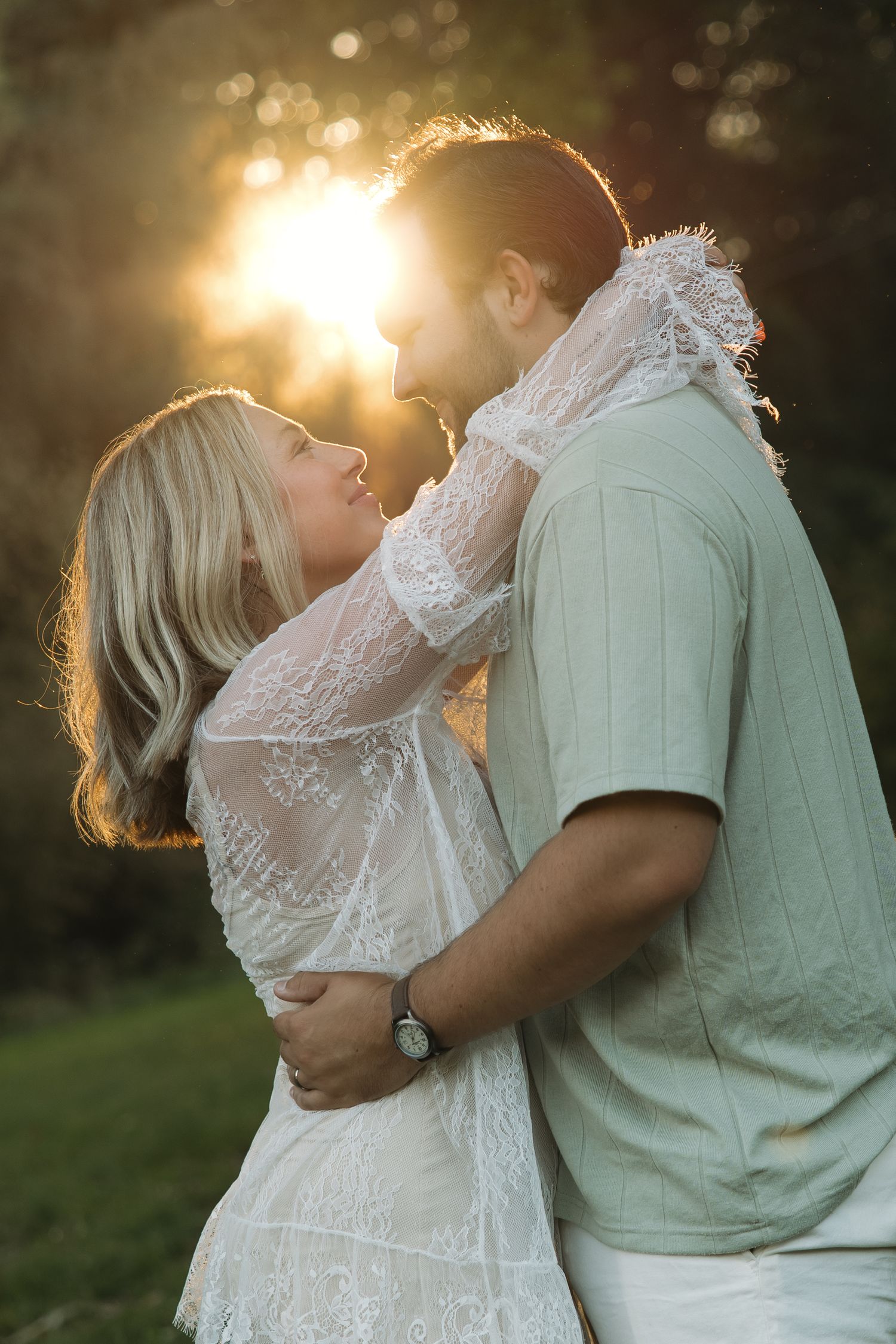 Couple embracing and smiling at each other in golden sunset light, woman in white lace dress arms around mans neck.