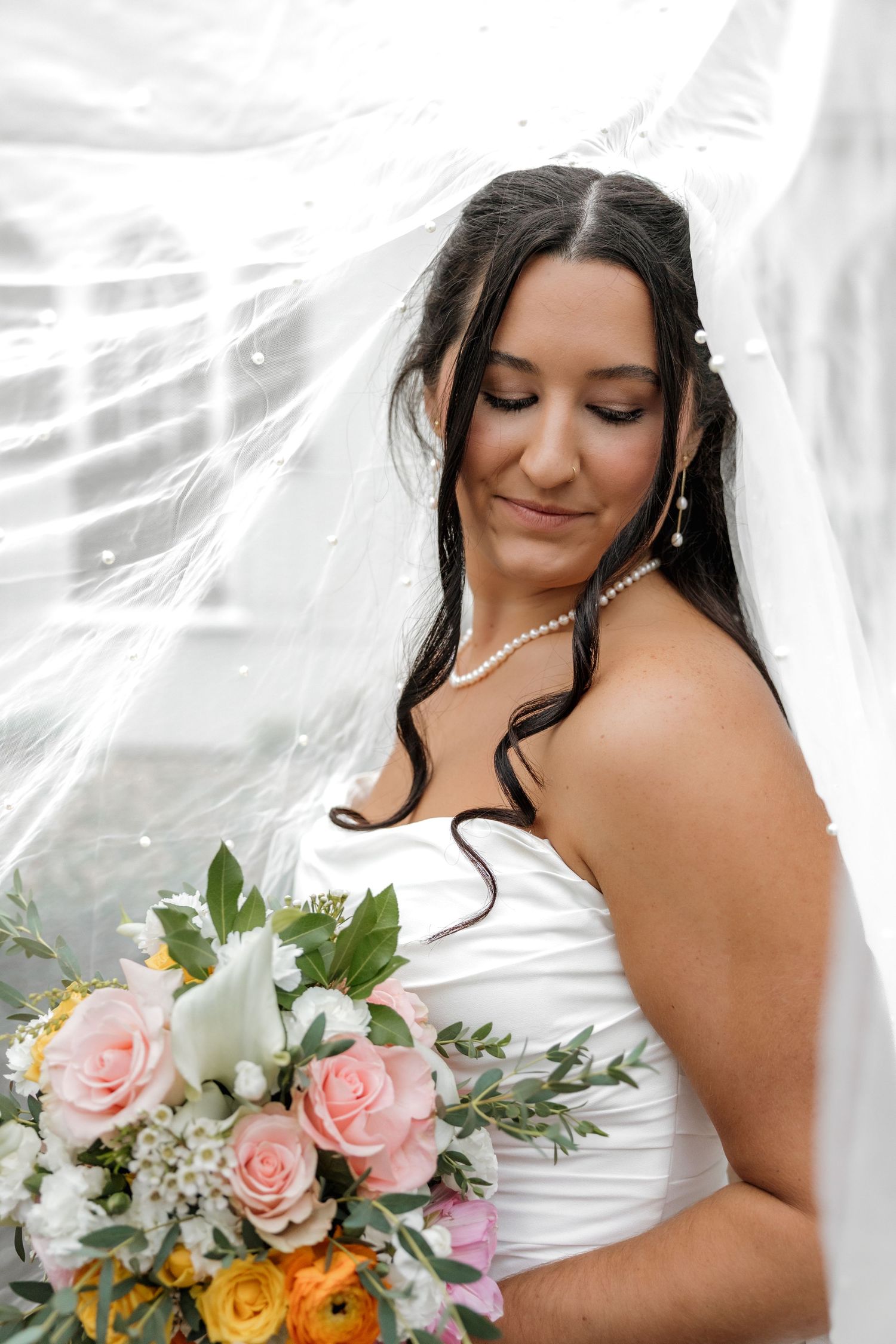 Beautiful dark-haired bride in a strapless white gown holding a colorful bouquet under a flowing veil.