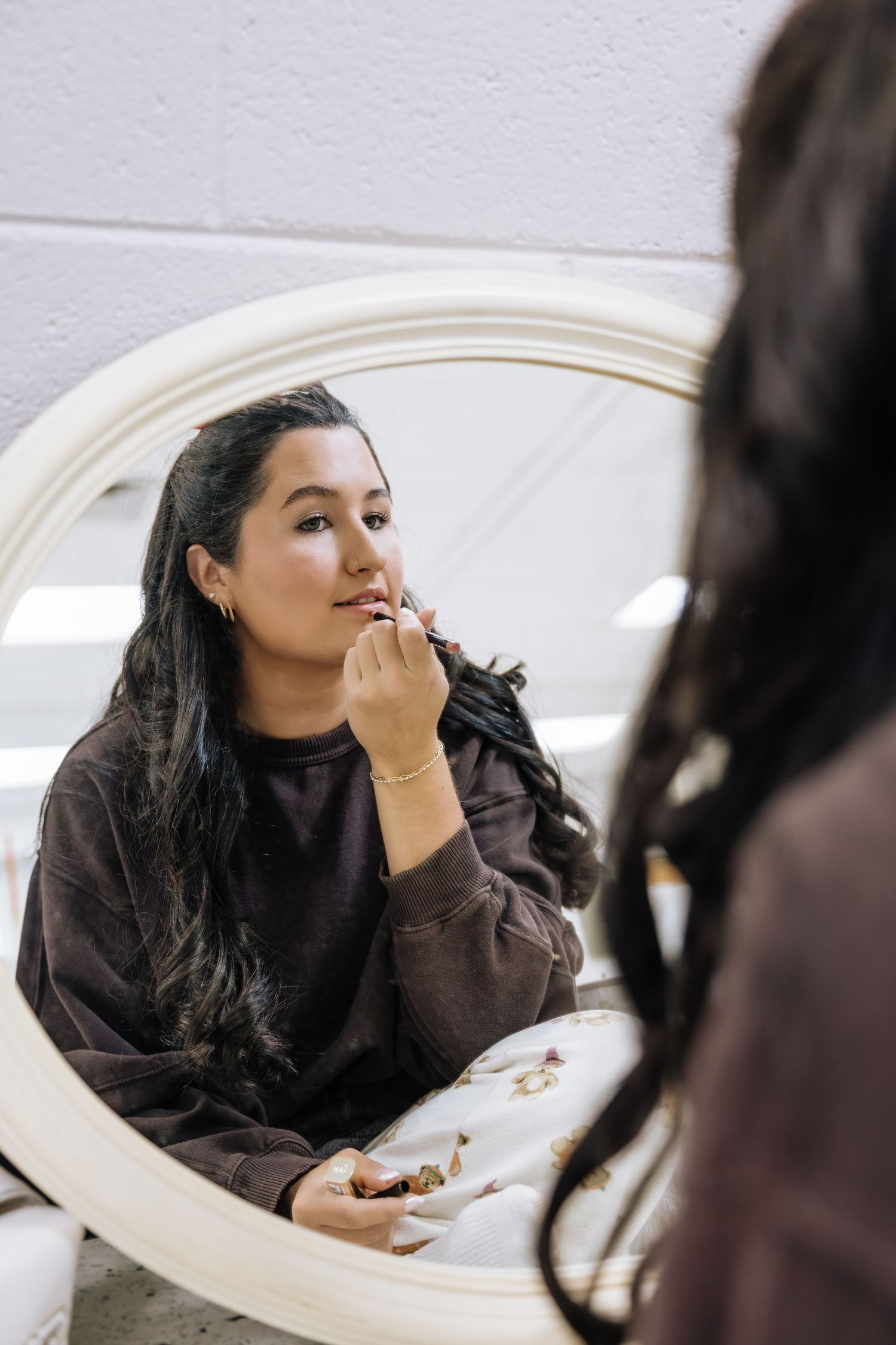 Bride applying lipstick while getting ready, reflected in round white vanity mirror.