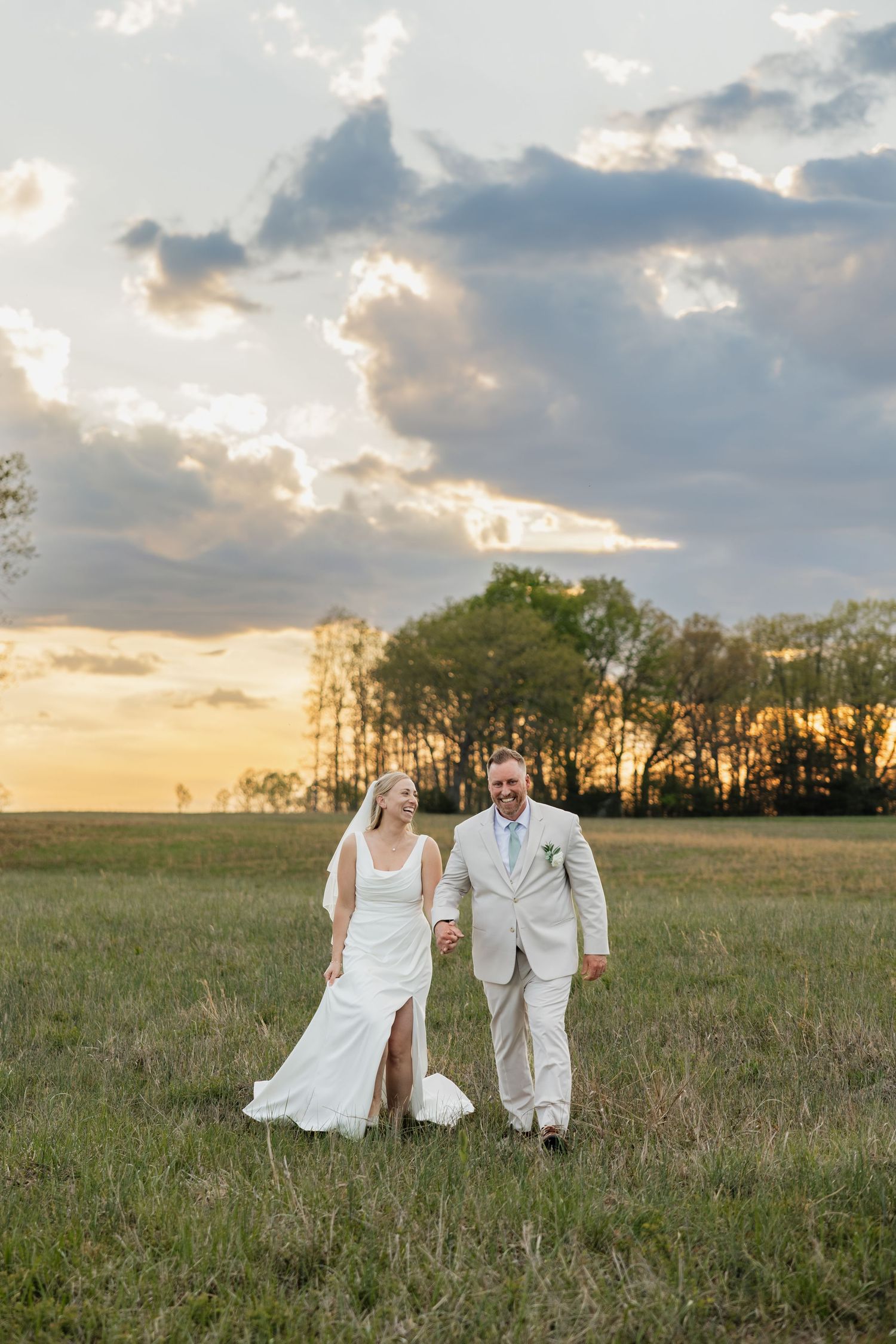 Bride and groom walk hand in hand through a green field at golden hour sunset.
