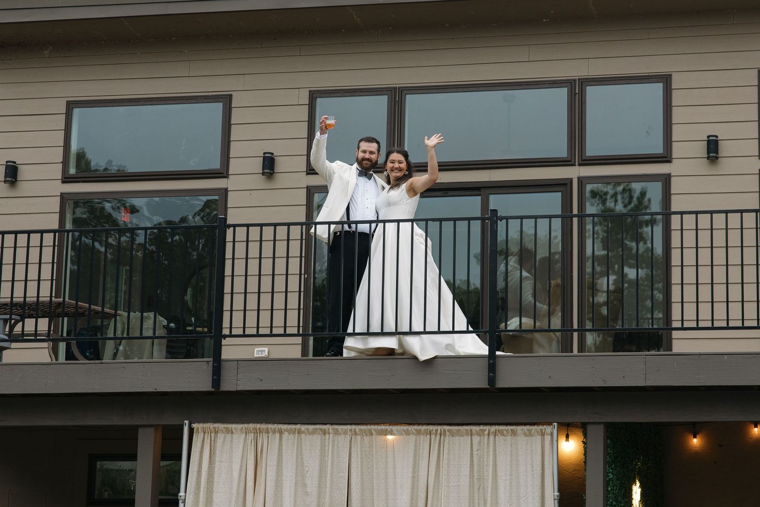 Excited bride and groom waving from a balcony of a modern wedding venue building.
