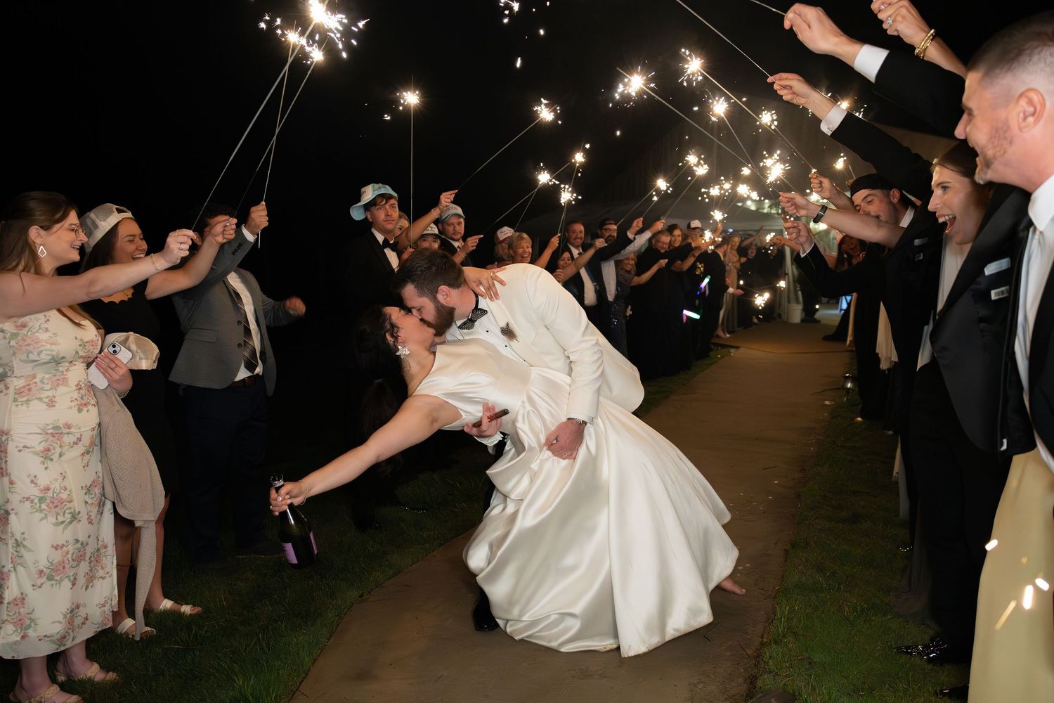 Bride and groom share romantic dip kiss during sparkler send-off exit surrounded by cheering wedding guests at night.