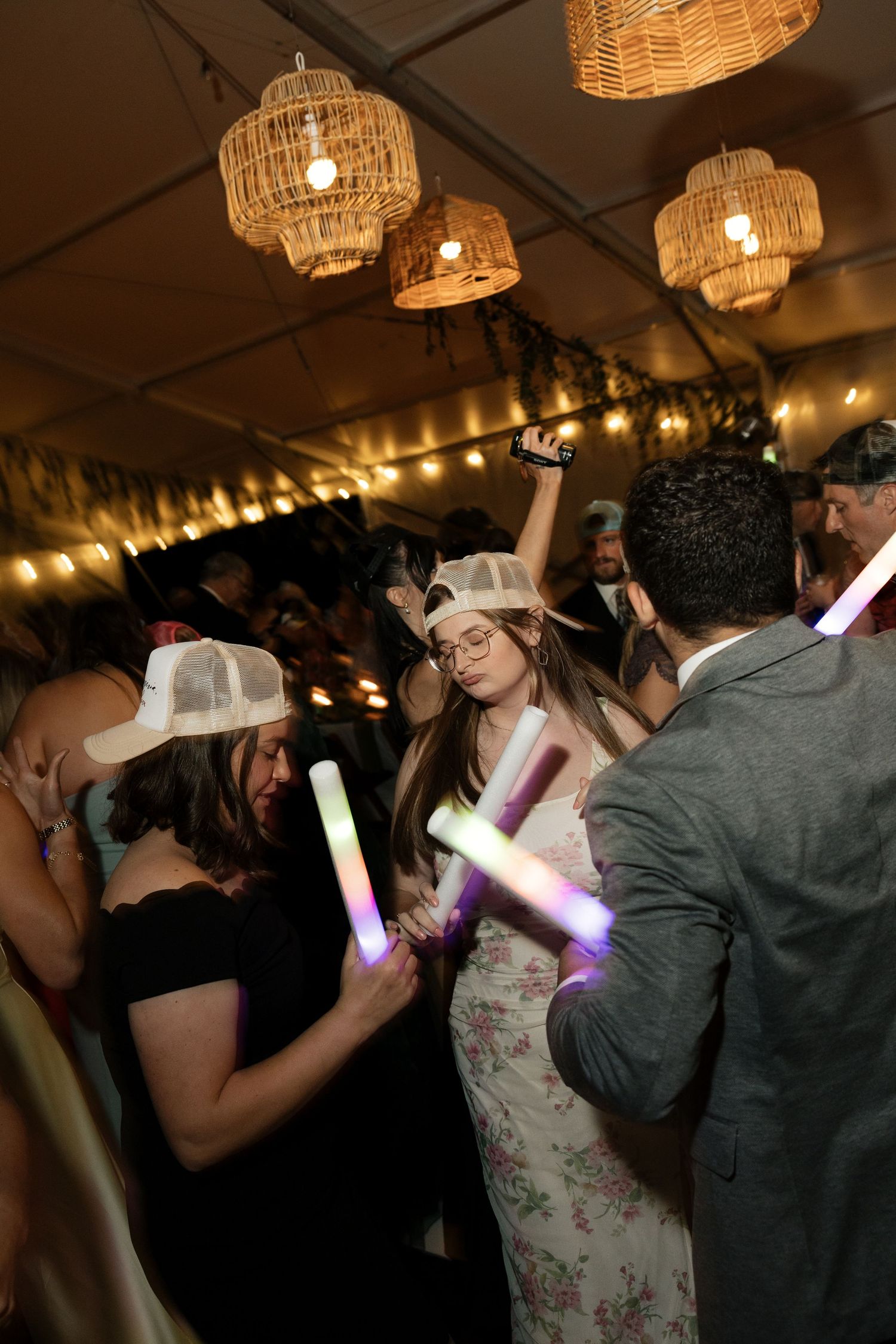 Guests dancing at a wedding reception under wicker pendant lights, holding colorful LED foam glow sticks.